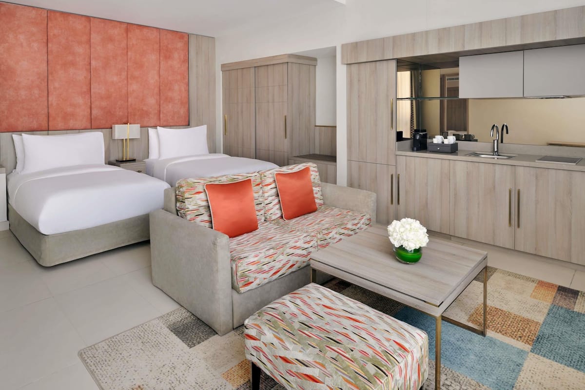 A modern hotel room with two beds, a sofa, a small table, and a kitchenette.
