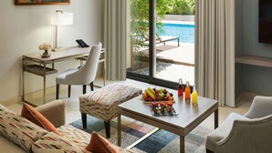 Habitación de hotel con escritorio, sofá, mesa de centro con fruta y bebidas, televisor y vista a una piscina.
