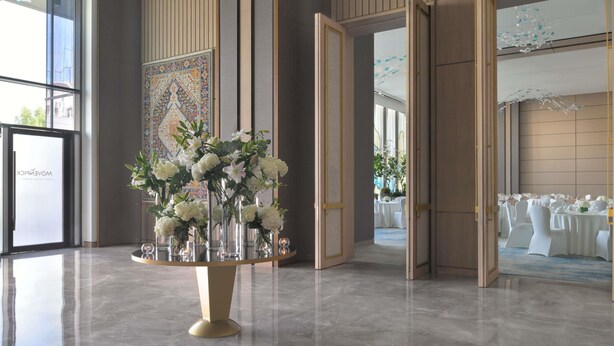 Un vestíbulo espacioso con una mesa redonda central decorada con flores blancas, un gran tapiz con motivos en la pared y vistas a un salón de banquetes.