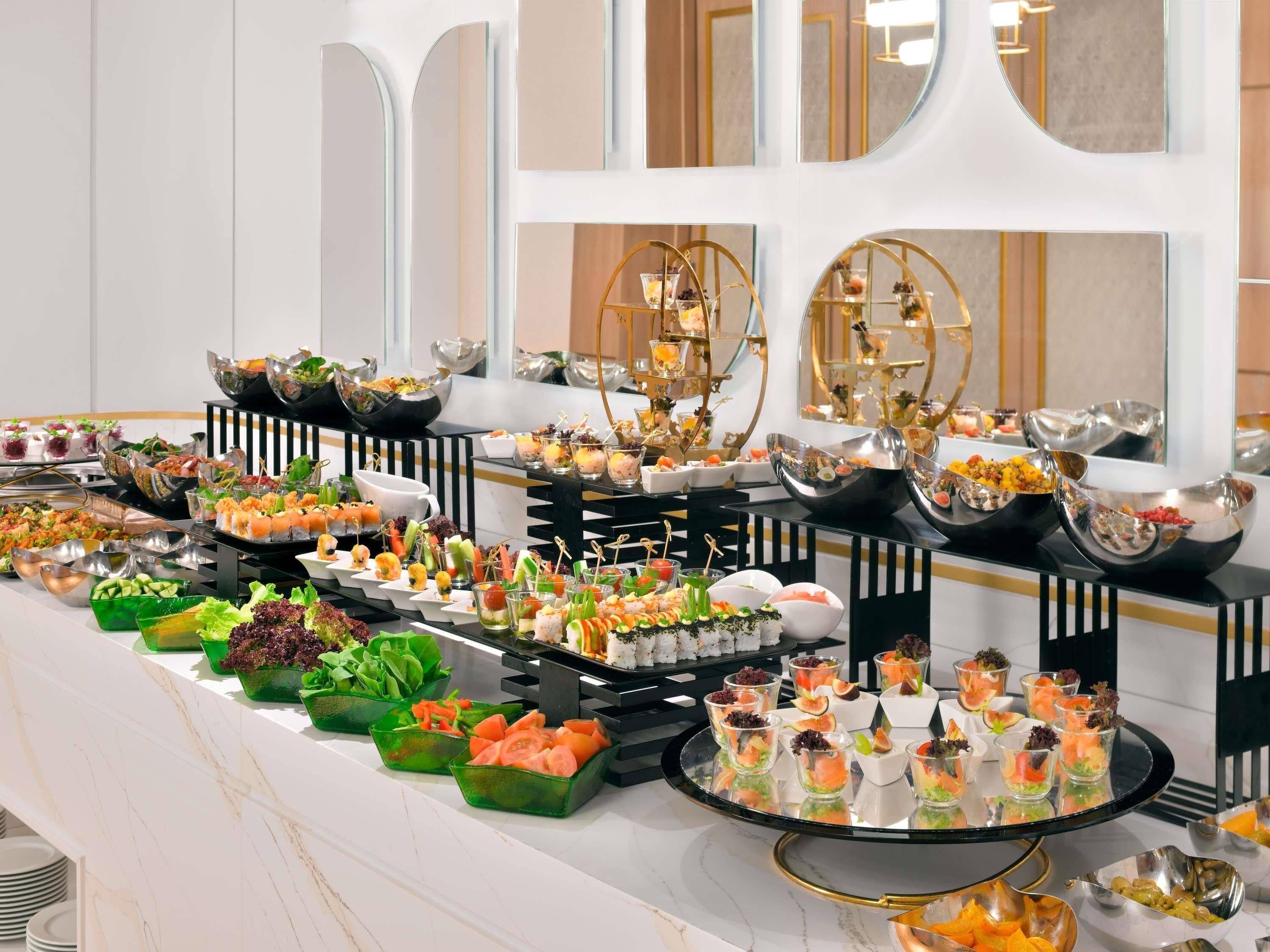 Un bufé con diversos alimentos y bebidas, que incluye ensaladas, aperitivos y postres, dispuesto sobre una encimera de mármol.