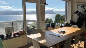 A room with a large window overlooking a body of water and mountains, a wooden table, and a hanging chair. - Guesthouse Stekkaból (Vesturbyggð)
