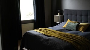 Une chambre à coucher avec un lit de couleur sombre, une table de chevet avec une lampe et une fenêtre avec des rideaux.