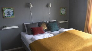 Un lit bien fait, recouvert d’un plaid jaune, agrémenté de plusieurs coussins de différentes couleurs et éclairé par deux lampes fixées au mur.
