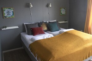Un lit bien fait, recouvert d’un plaid jaune, agrémenté de plusieurs coussins de différentes couleurs et éclairé par deux lampes fixées au mur.