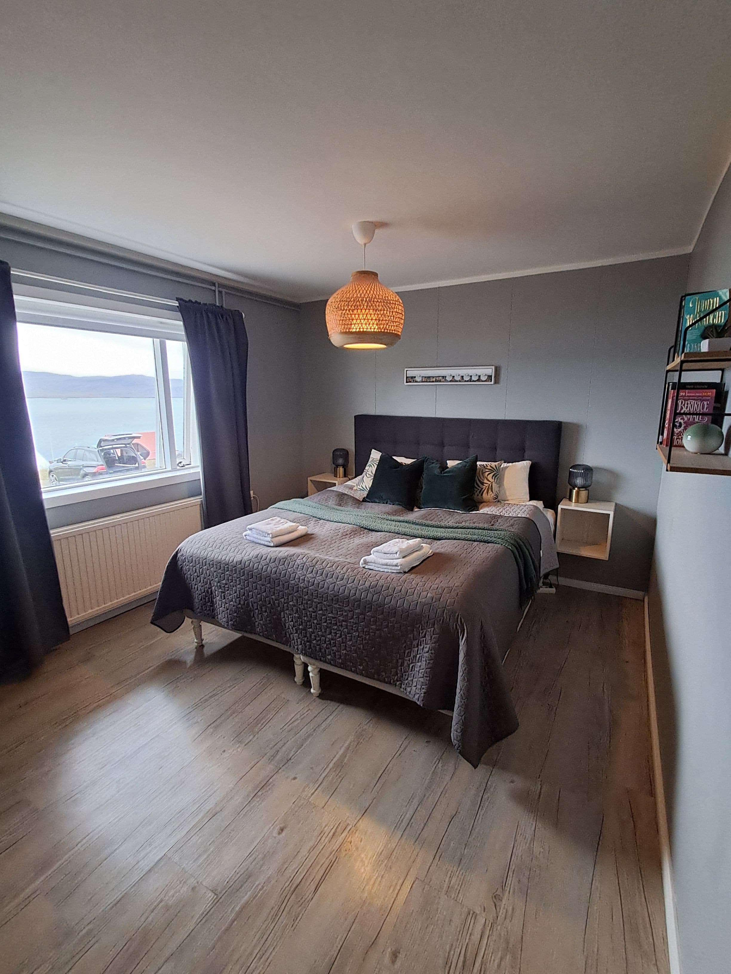Een slaapkamer met een groot bed, nachtkastjes, een hanglamp, uitzicht op zee en een raam met gordijnen.