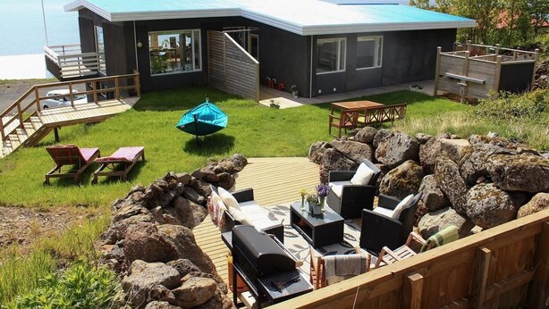A modern house with a deck, outdoor seating, and a view of the water. - Guesthouse Stekkaból (Vesturbyggð)