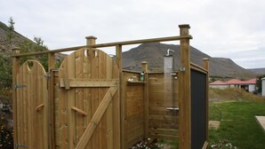 A wooden outdoor shower enclosure with a metal showerhead, surrounded by a wooden deck and a stone pathway. - Guesthouse Stekkaból (Vesturbyggð)