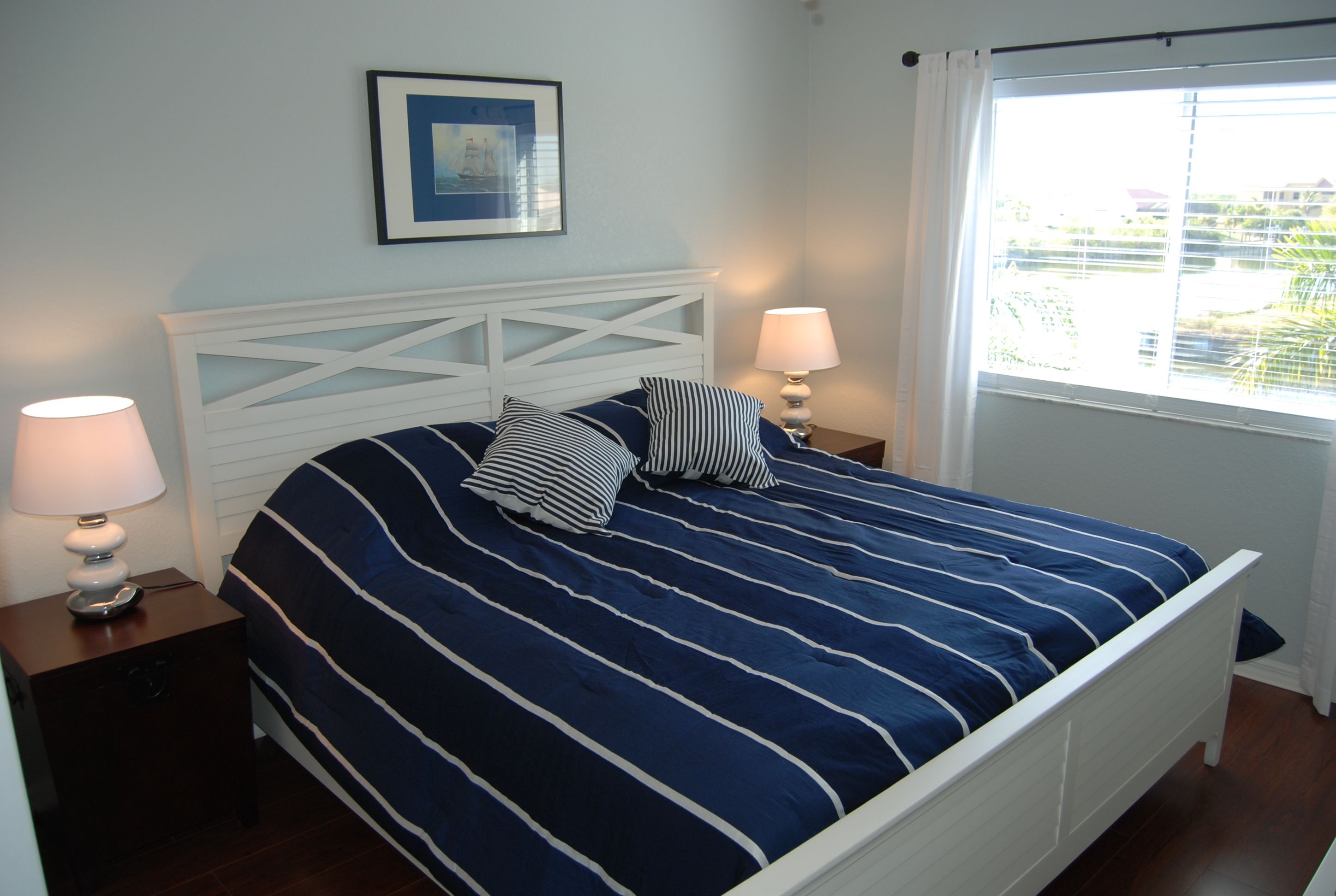 A neatly made bed with a striped blue and white bedspread, two pillows, and a white headboard. There is a framed picture on the wall above the bed and a window with blinds to the right.