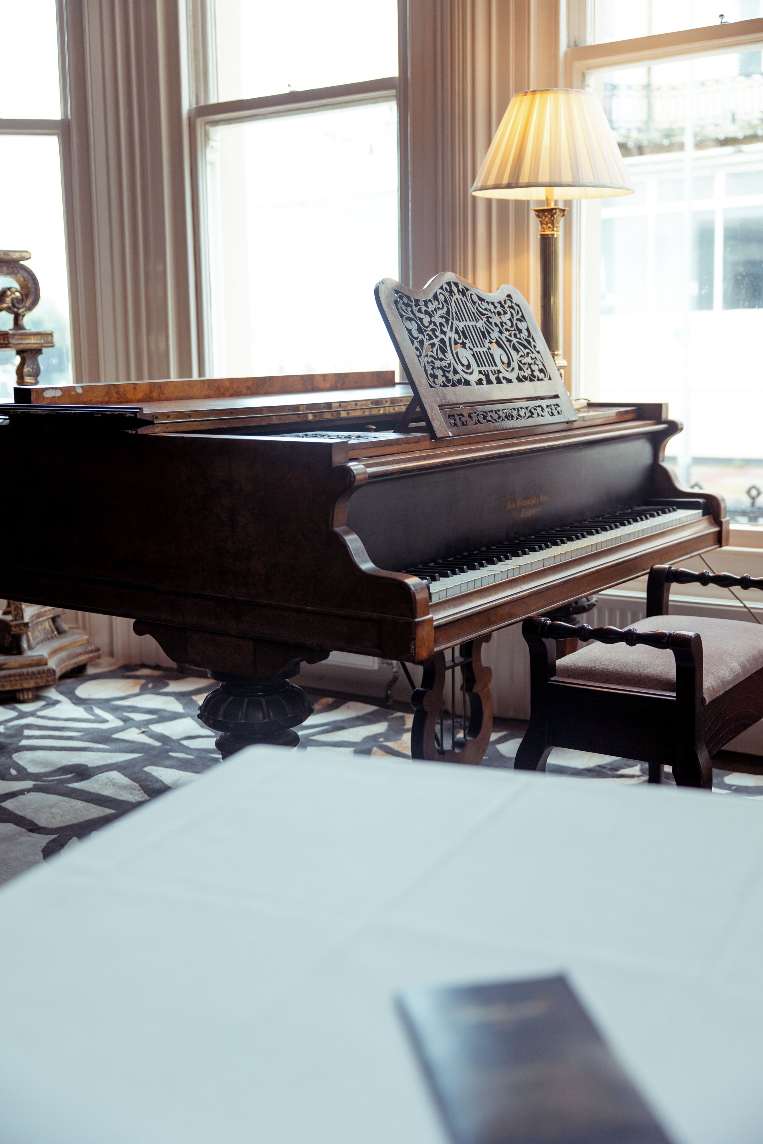 A grand piano in a well-lit room with large windows, a patterned floor, and a lamp.
