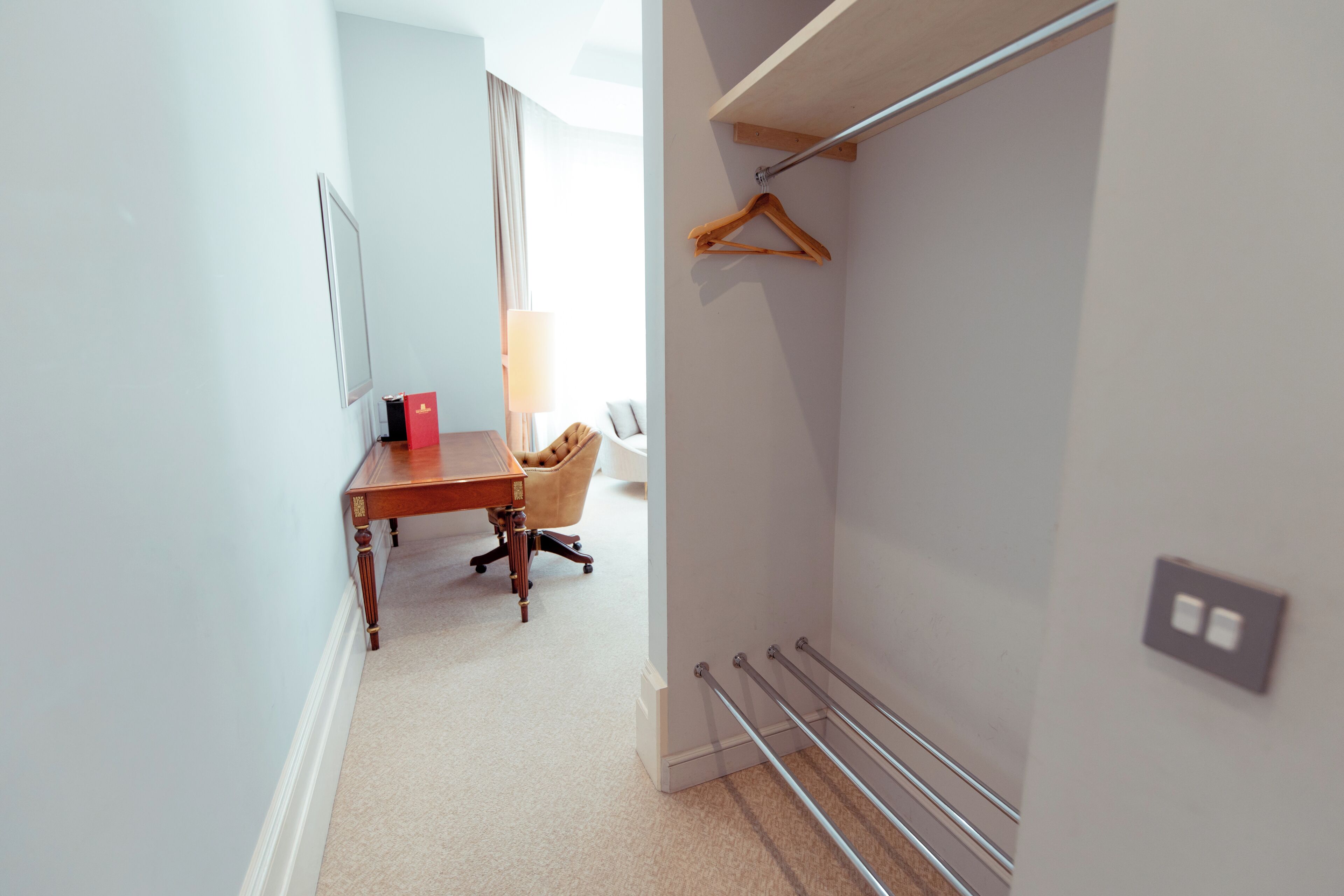 A hotel room with a wooden desk, a chair, a hanging coat rack, and a bedside table.