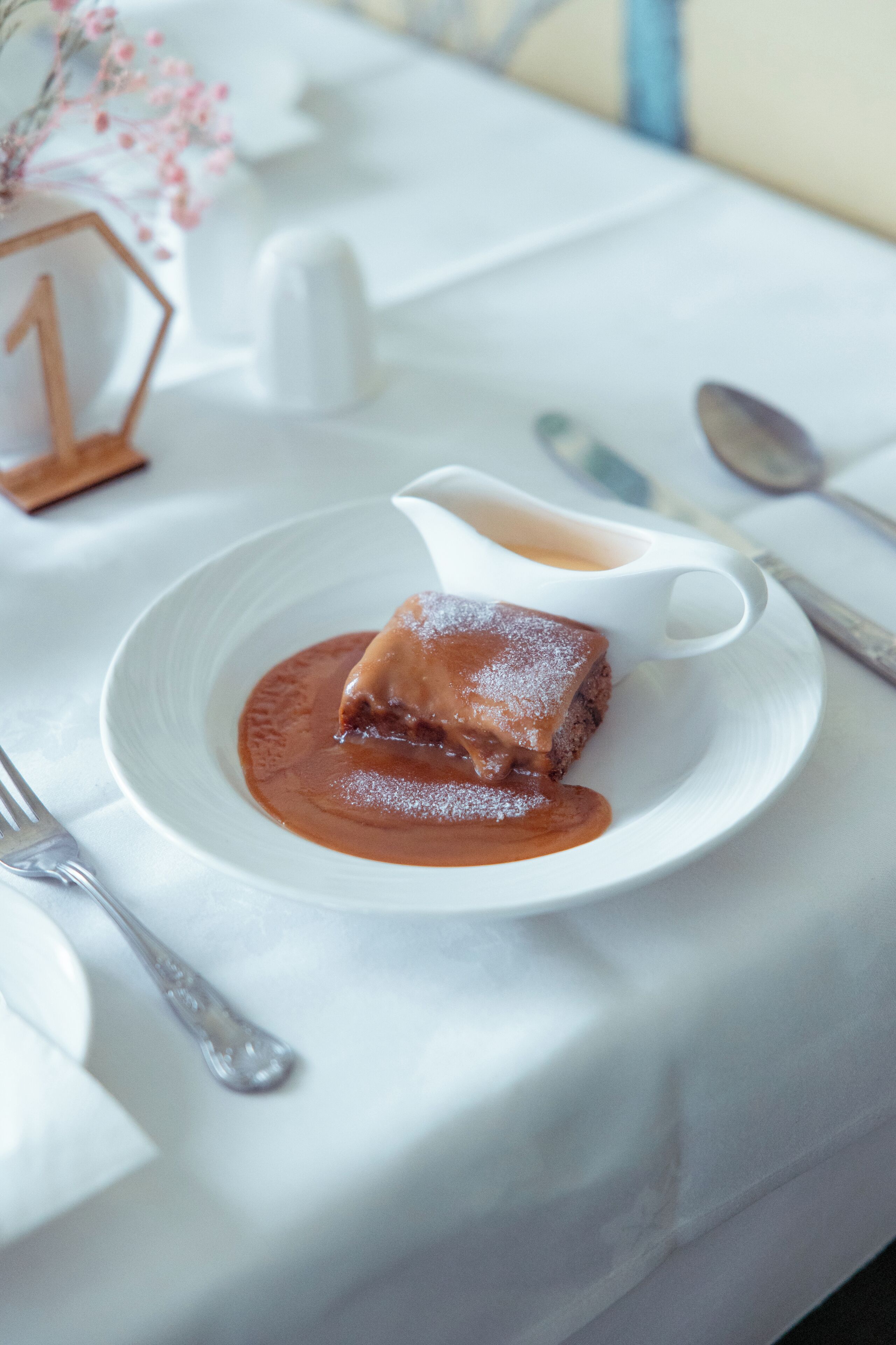 A dessert on a white plate with a sauce, a spoon, and a fork on a table with a white tablecloth.