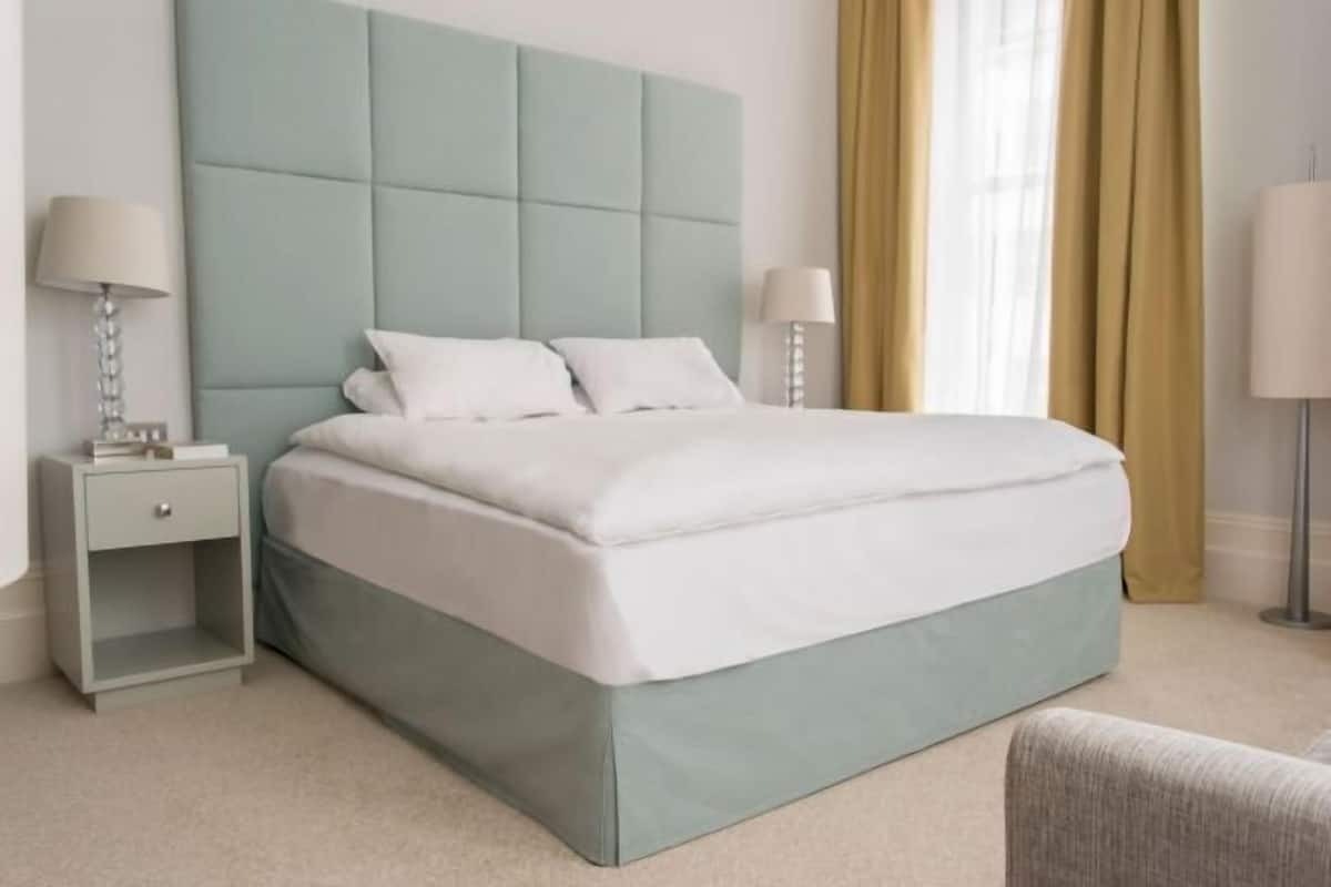 A neatly made bed with a light blue headboard, a bedside table with a lamp, and a chair.