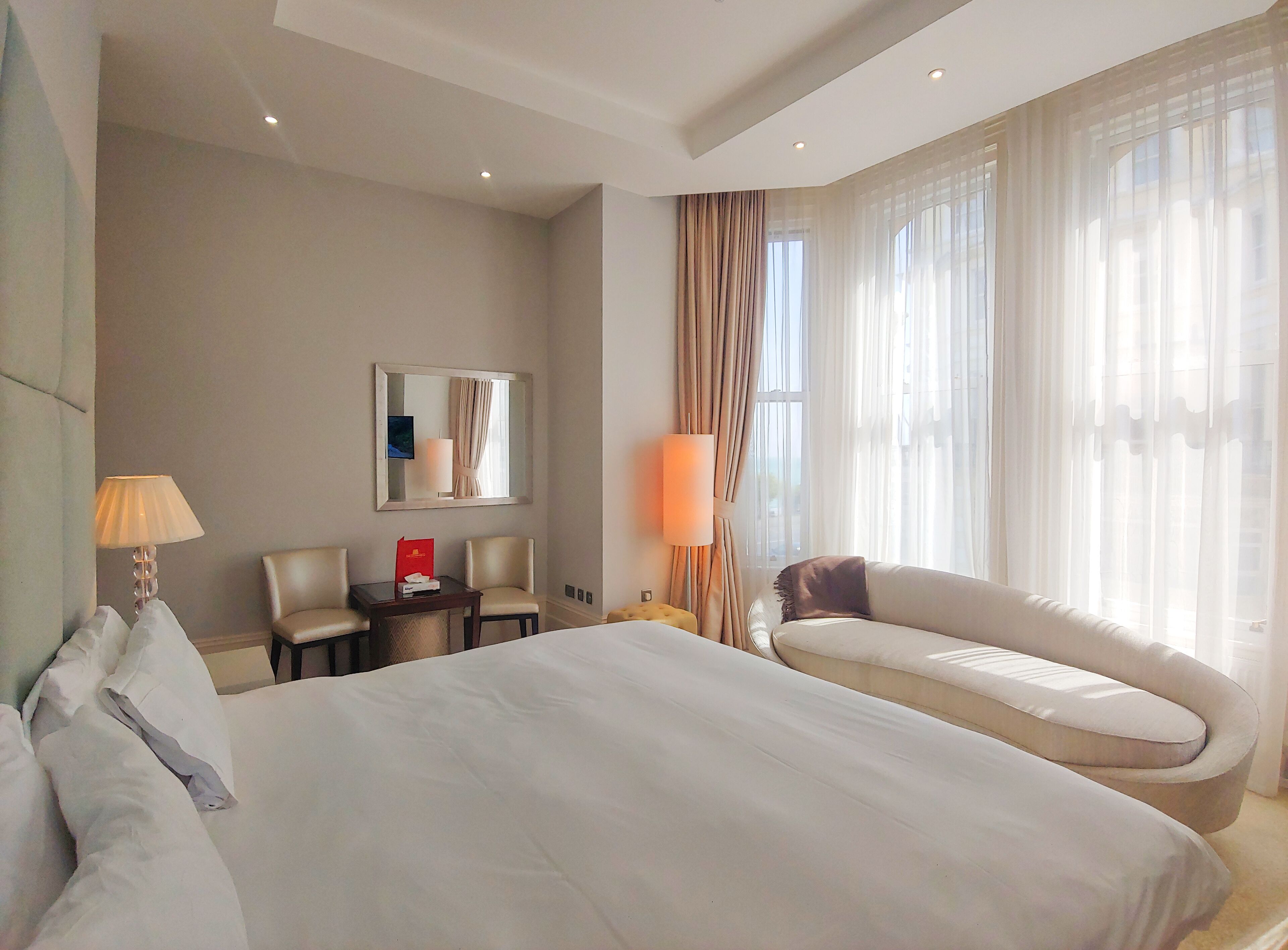 A hotel room with a large bed, two armchairs, a sofa, a desk with a lamp, a mirror, and a window with curtains.