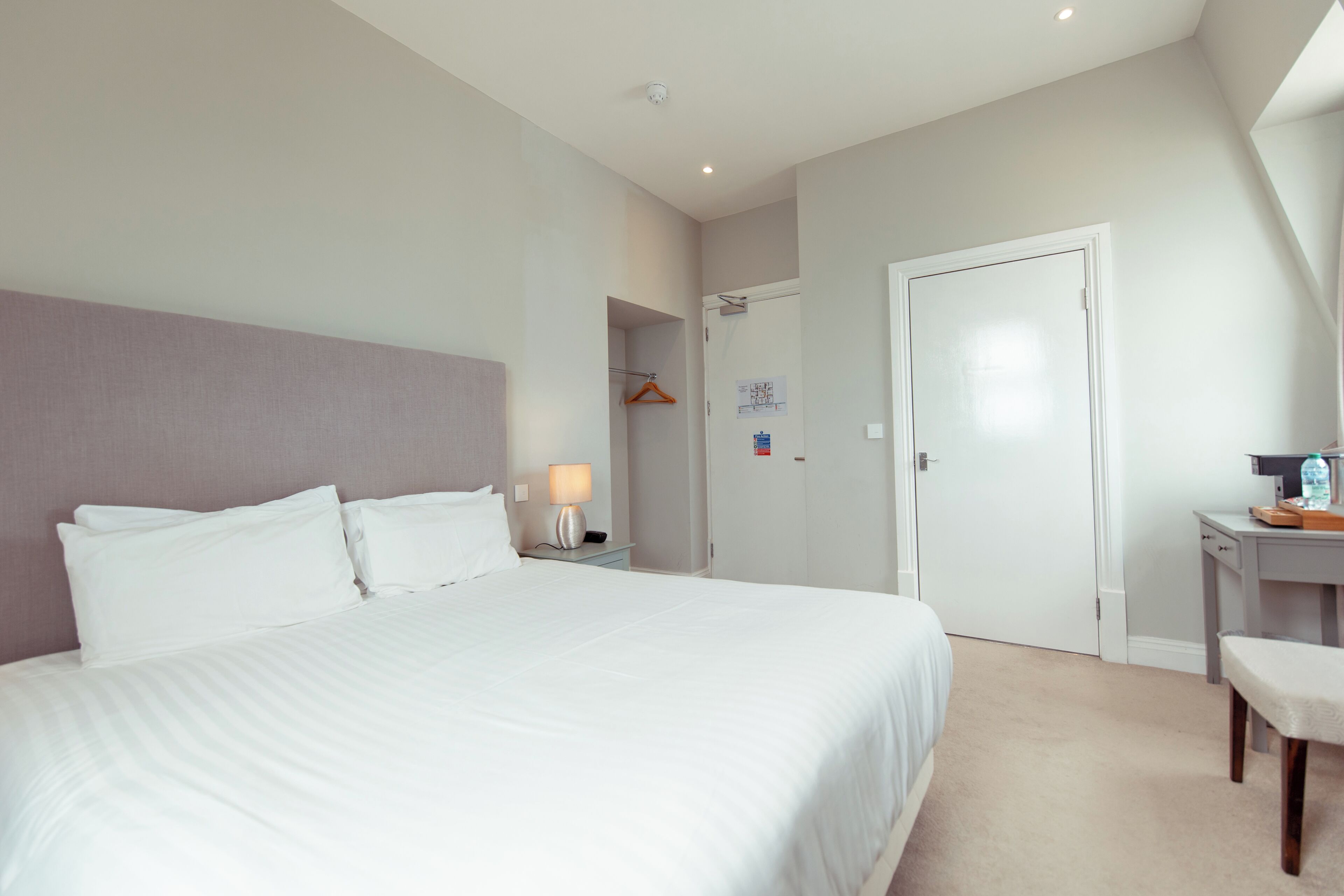 A hotel room with a large bed, a bedside table with a lamp, a desk with a chair, and a wardrobe.