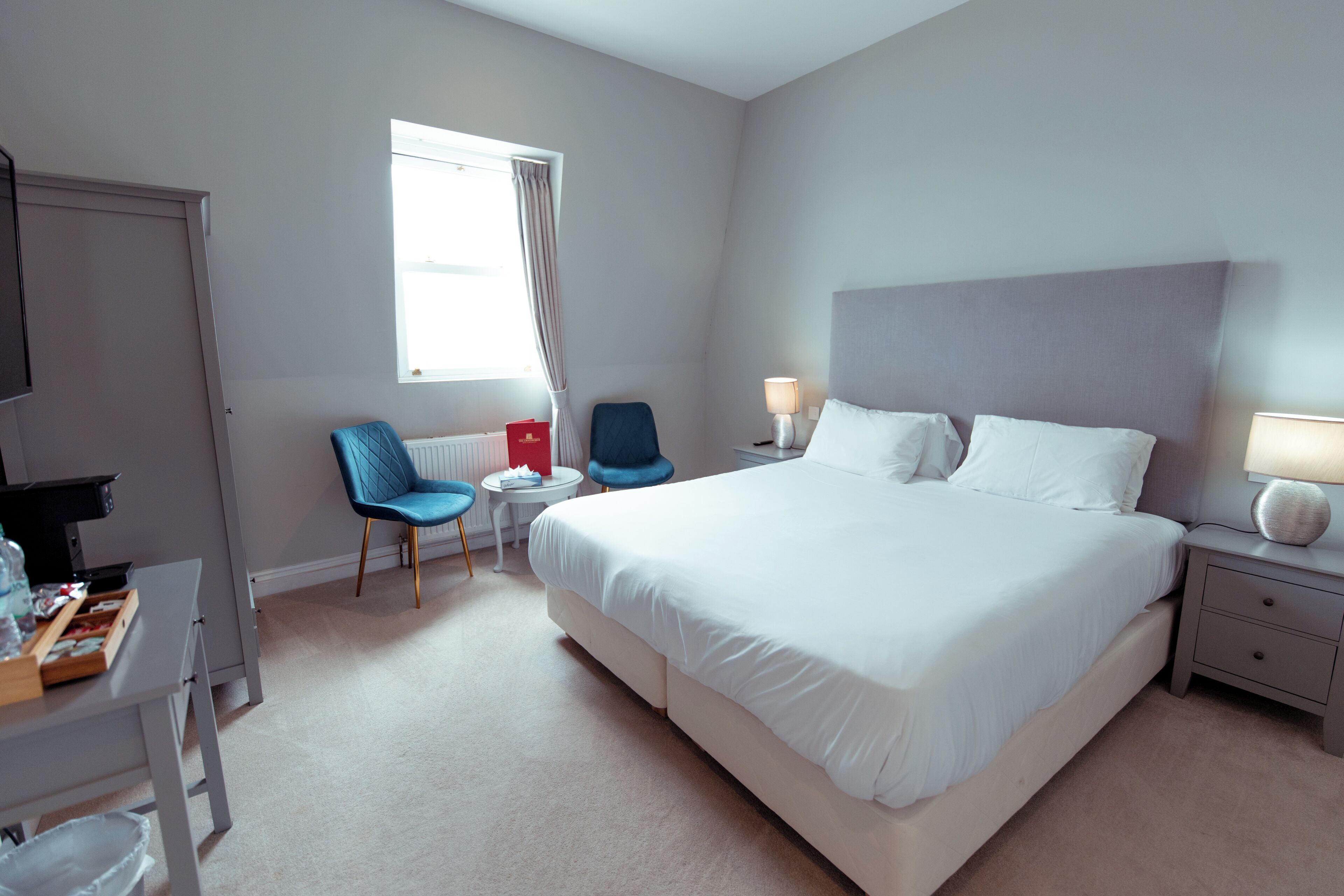 A hotel room with a large bed, a desk with a chair, a small table, and a window with curtains.