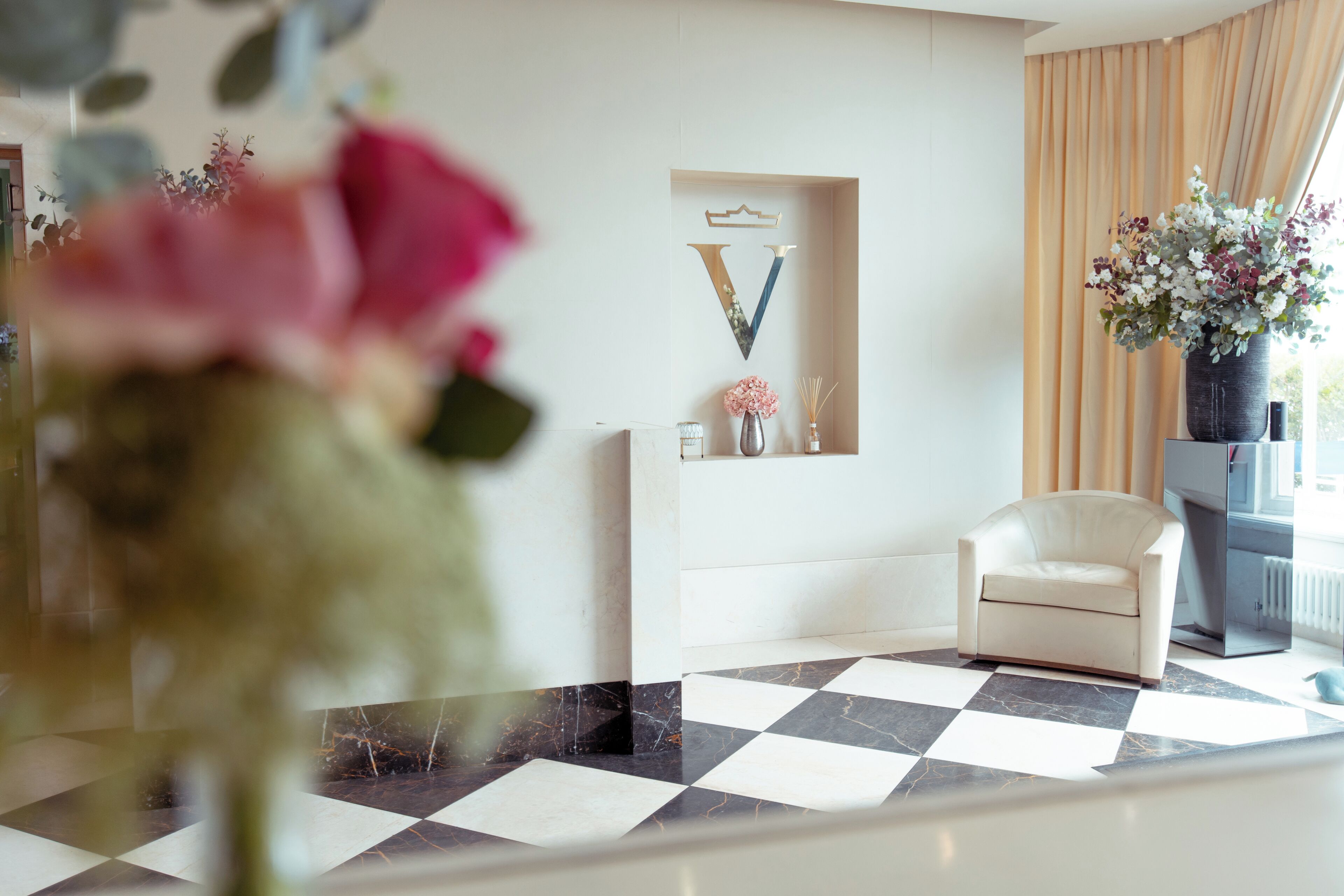 A modern hotel lobby with a checkered floor, a white armchair, a vase with flowers, and decorative wall art.