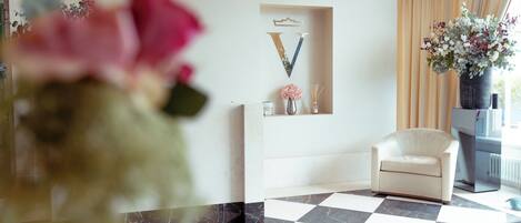 A modern hotel lobby with a checkered floor, a white armchair, a vase with flowers, and decorative wall art.