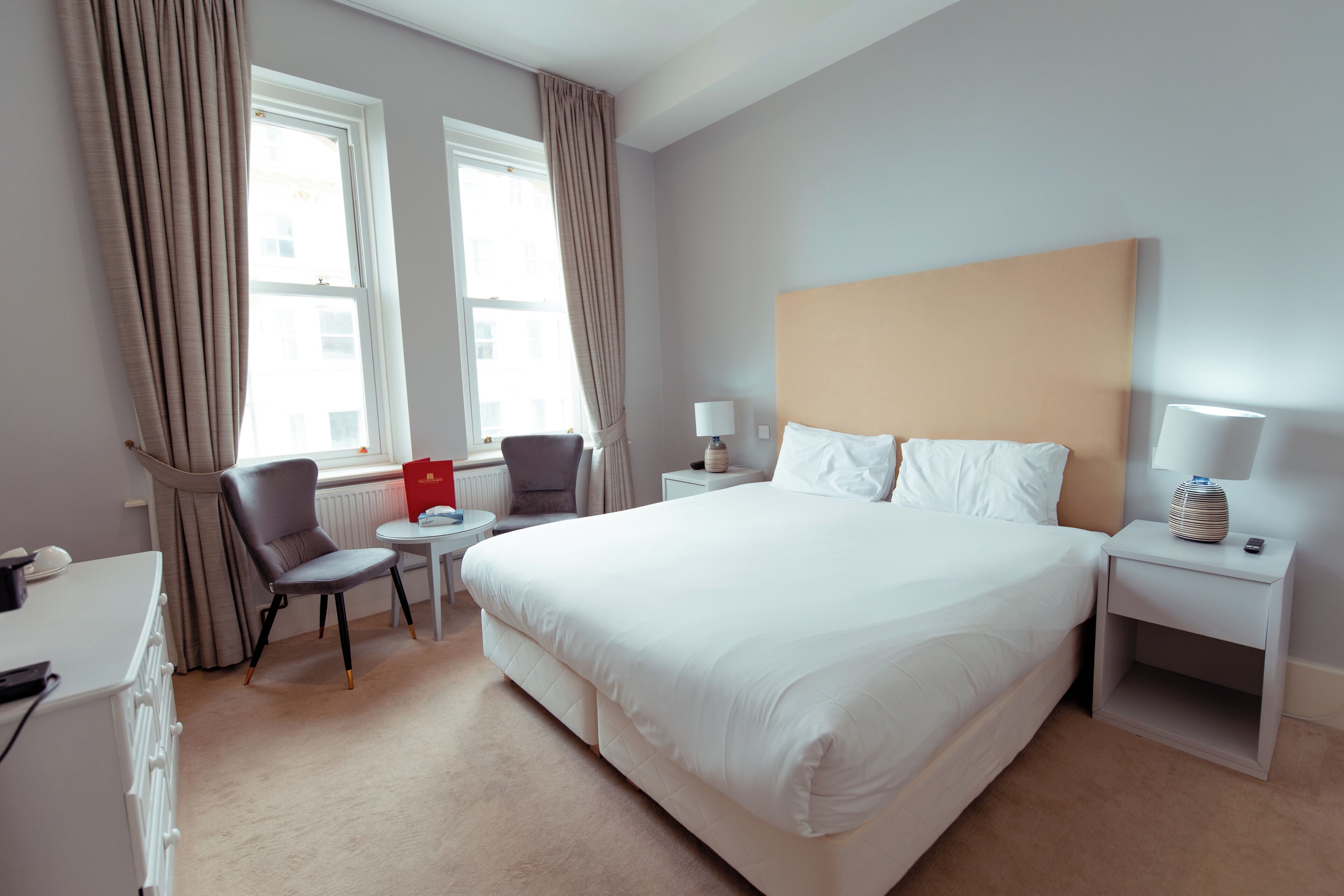 A hotel room with a large bed, a desk with a chair, a small table, and a nightstand with a lamp.