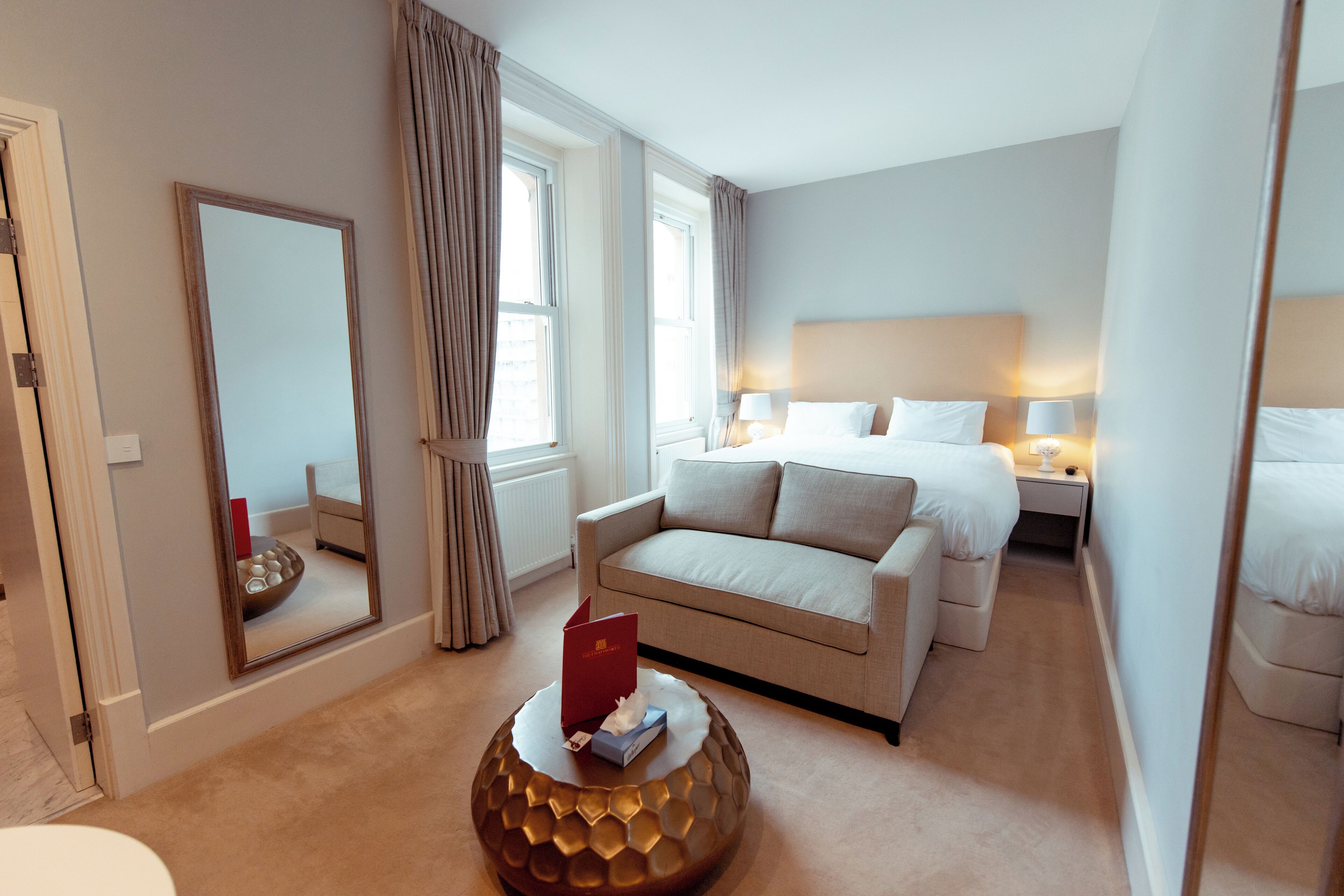 Habitación de hotel con cama, sofá, mesa de centro redonda, espejo y ventana con cortinas.