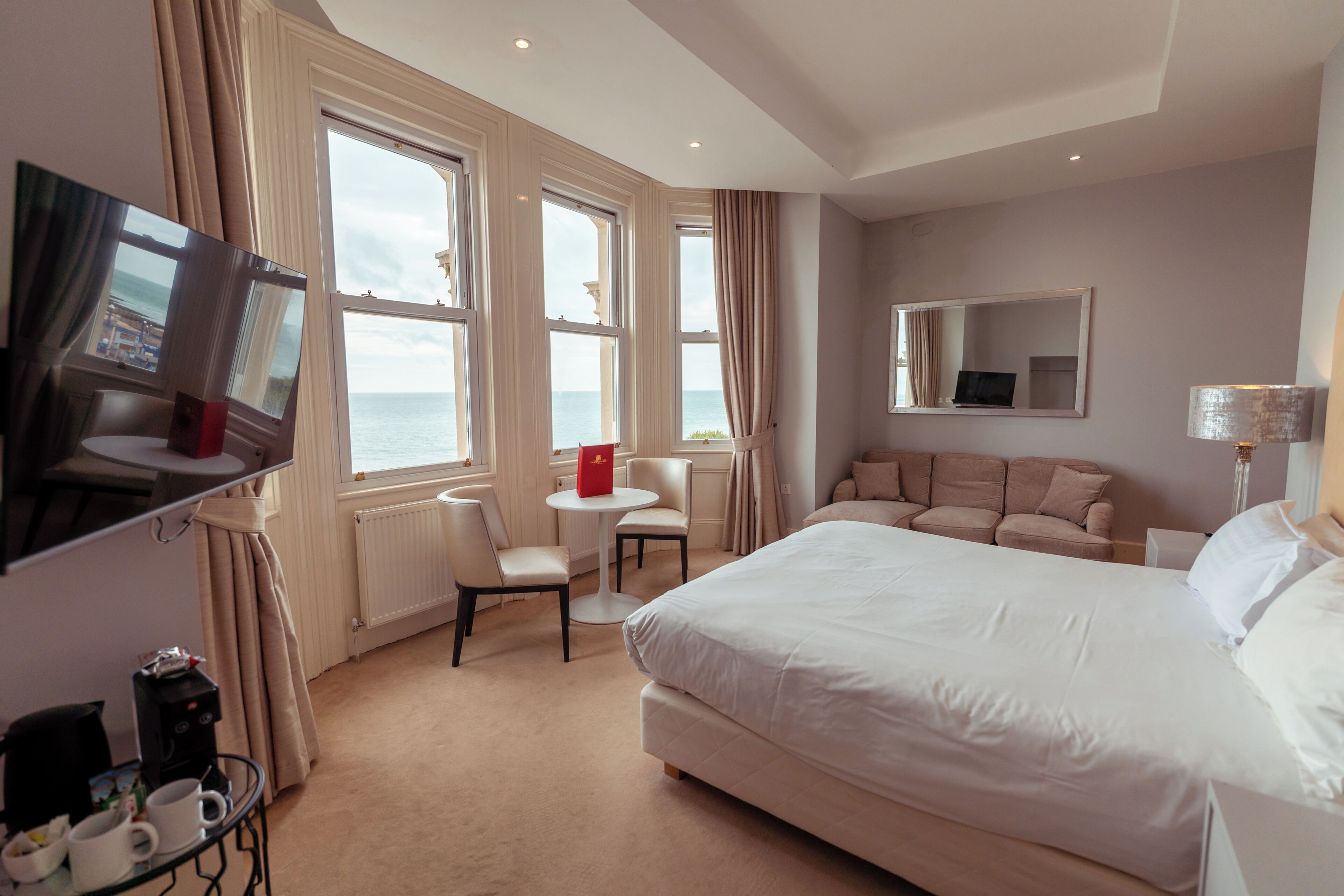 A hotel room with a large bed, a TV, a small table with chairs, a sofa, and a view of the sea through large windows.