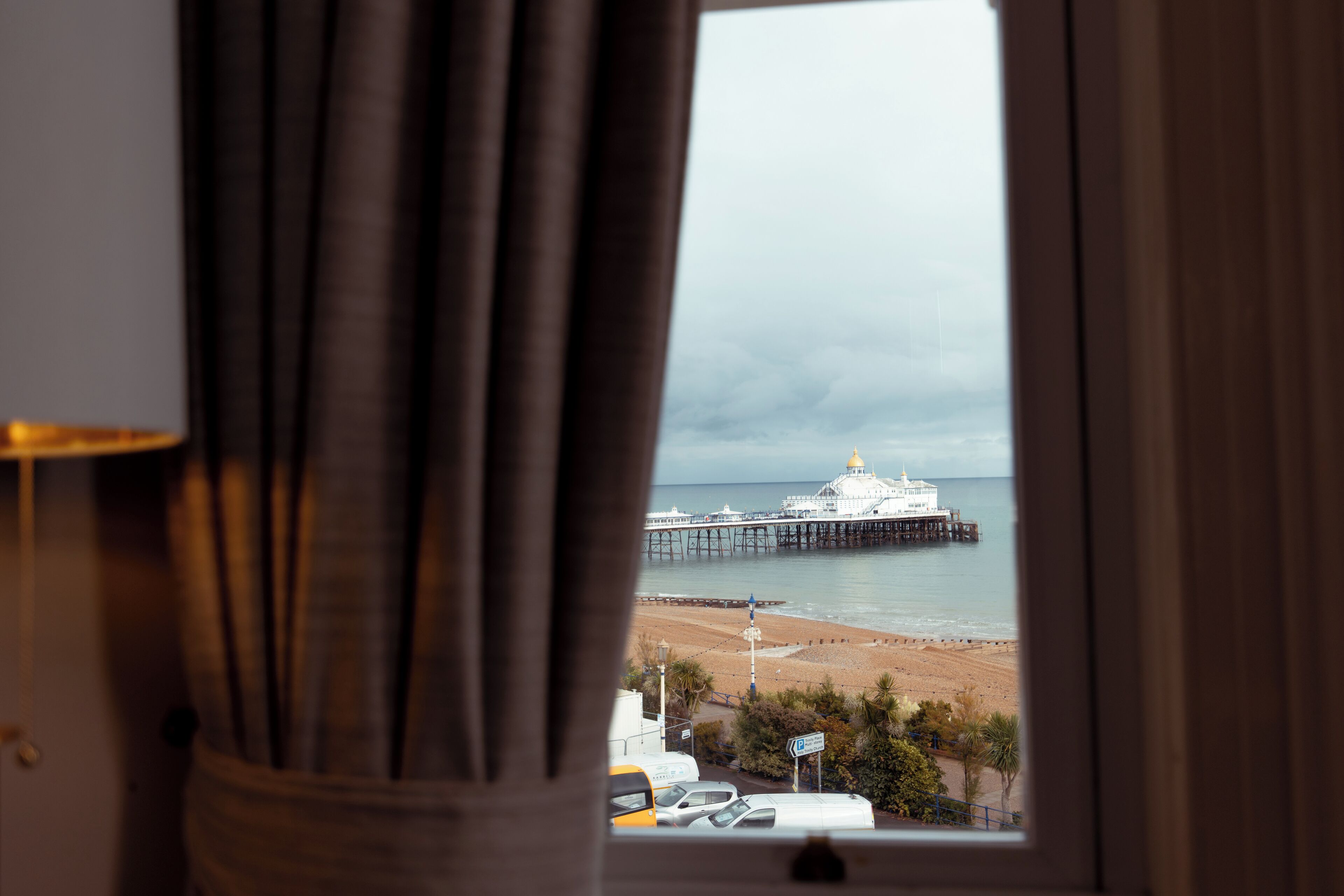 A view through a window with curtains, featuring a beach, a pier, and a white building with a dome.