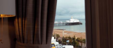 A view through a window with curtains, featuring a beach, a pier, and a white building with a dome.