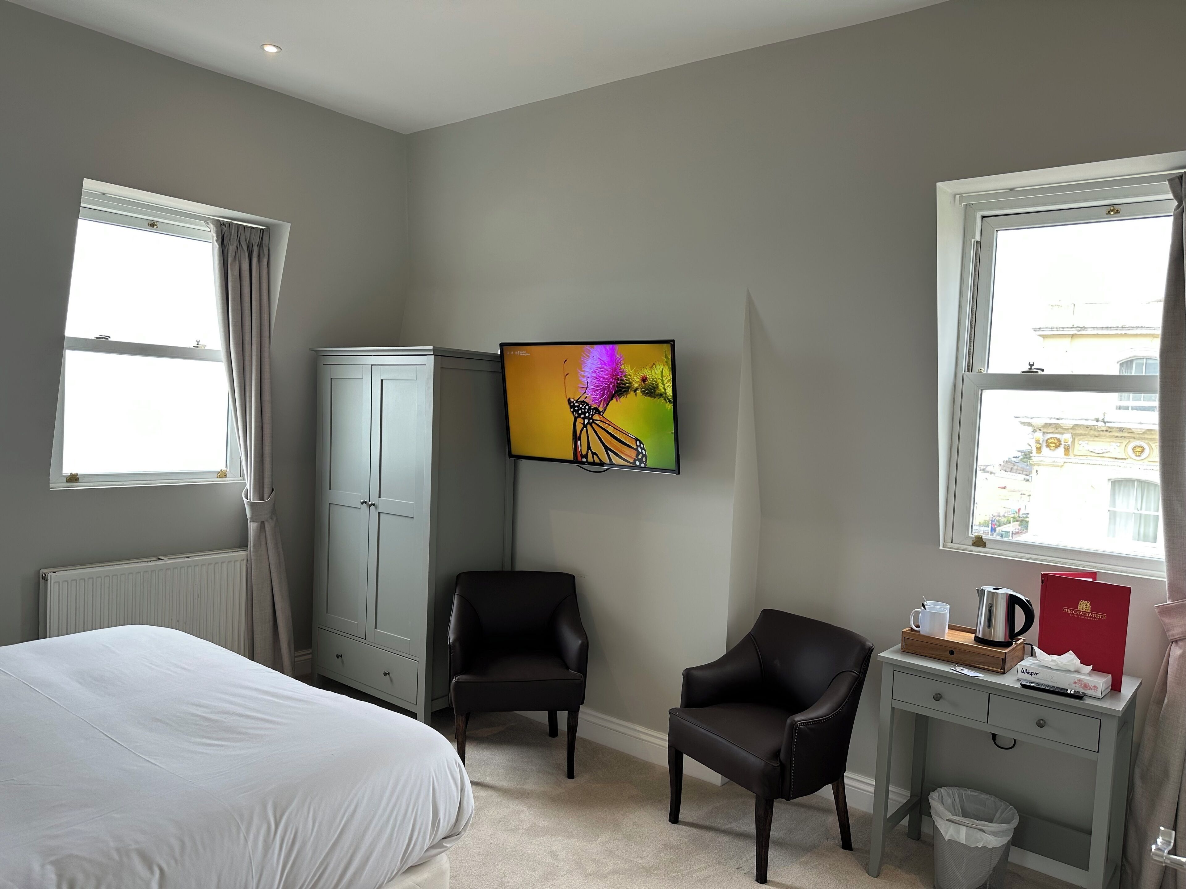 A hotel room with a bed, two chairs, a desk with a coffee pot, a TV on the wall, and a radiator.