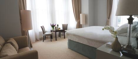 A hotel room with a bed, a desk, a chair, a lamp, and a vase with flowers.