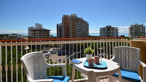 Un balcon avec des chaises en plastique blanches, une petite table sur laquelle se trouvent un pot de fleurs et une tasse, et une vue sur un parking et des bâtiments en arrière-plan.
