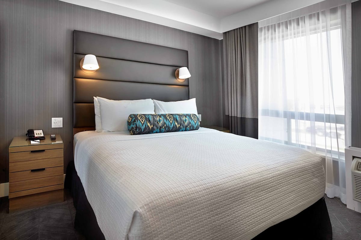 Ein modernes Hotelzimmer mit einem großen Bett, einem Nachttisch mit Lampe und Blick ins Freie durch transparente Vorhänge.