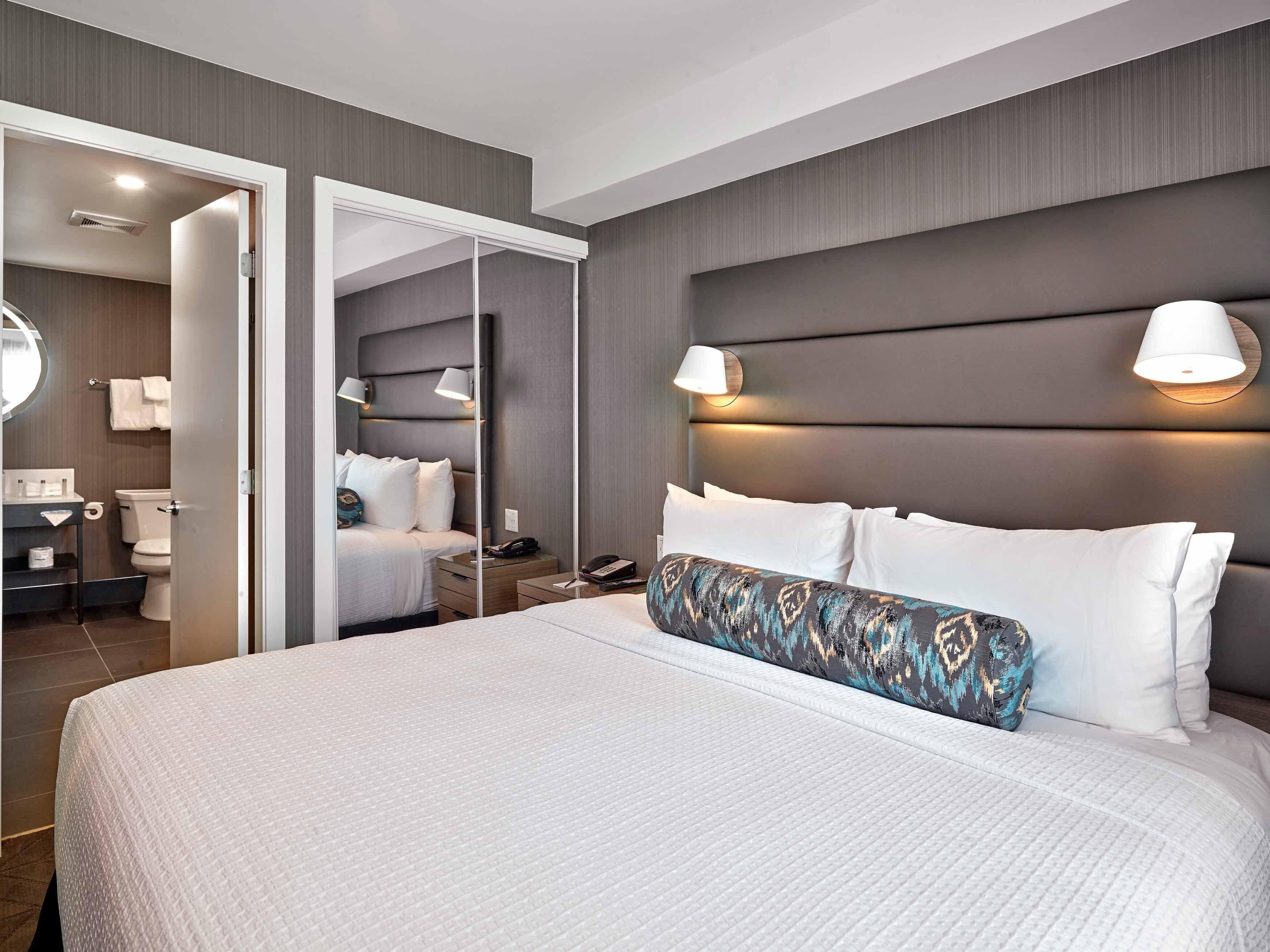 A hotel room with a large bed, a bedside table, a mirror, a nightstand, and a bathroom visible through an open door.