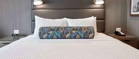 Habitación de hotel con una cama grande, dos lámparas de noche, un cojín estampado, una mesita de noche con un teléfono y un reloj de pared.