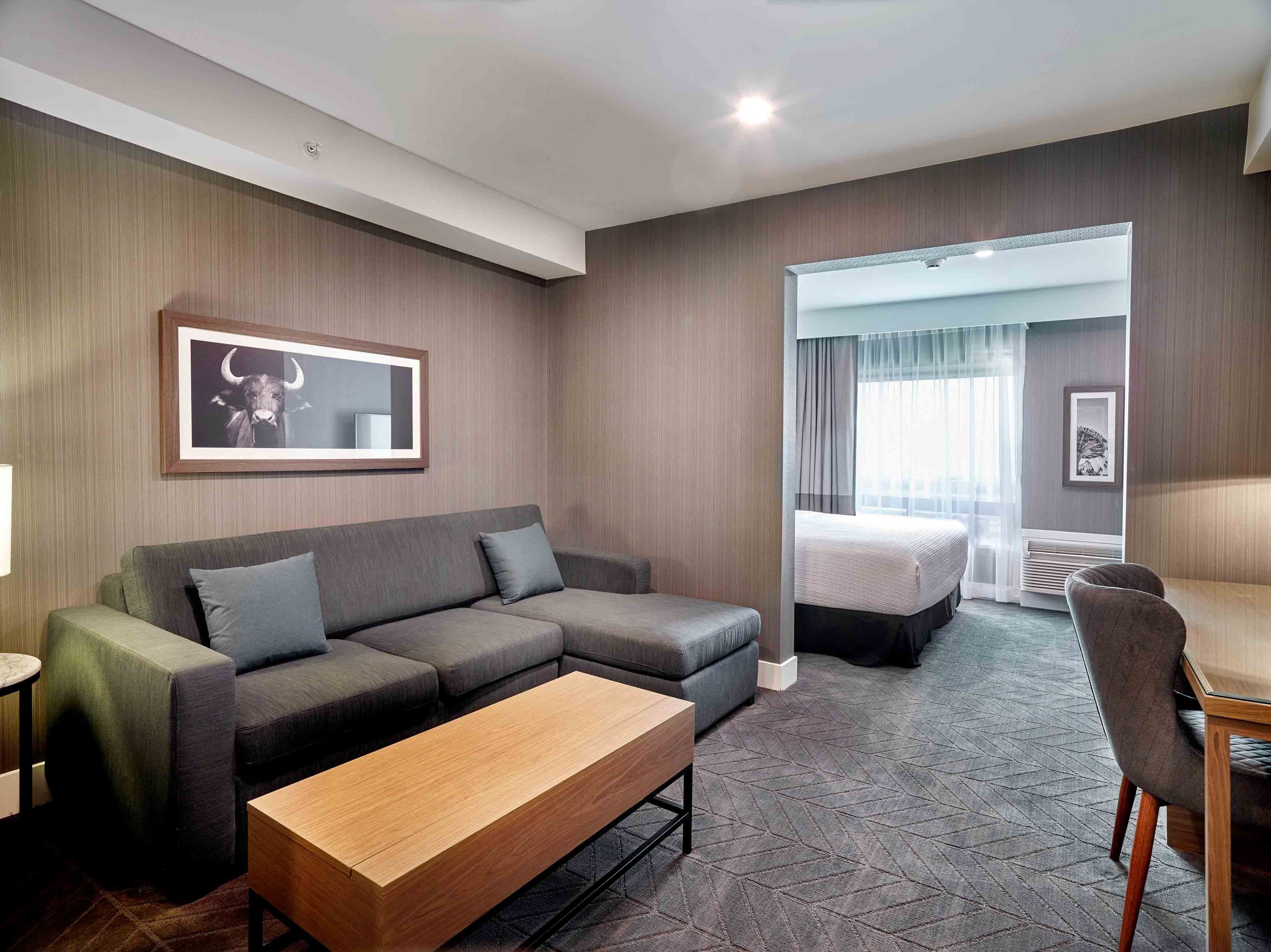 A hotel room with a grey sectional sofa, a wooden coffee table, a bed with a white headboard, and a small desk area.