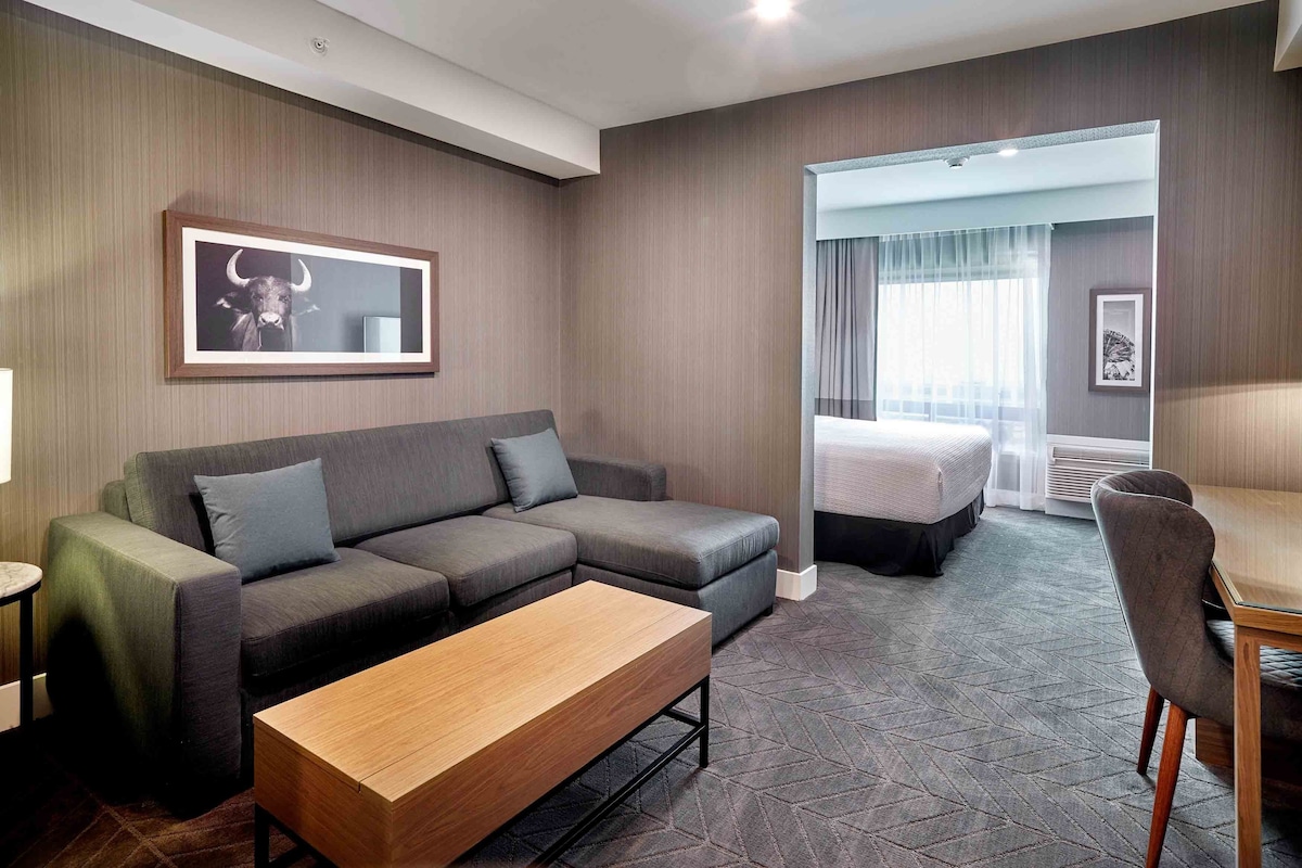 Une chambre d’hôtel avec un canapé d’angle gris, une table basse en bois, un lit avec une tête de lit blanche et un petit espace bureau.