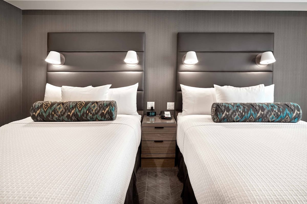 Ein Hotelzimmer mit zwei Betten, jedes mit einem gemusterten Kissen, einem Nachttisch mit einer Uhr und einer Wandlampe.