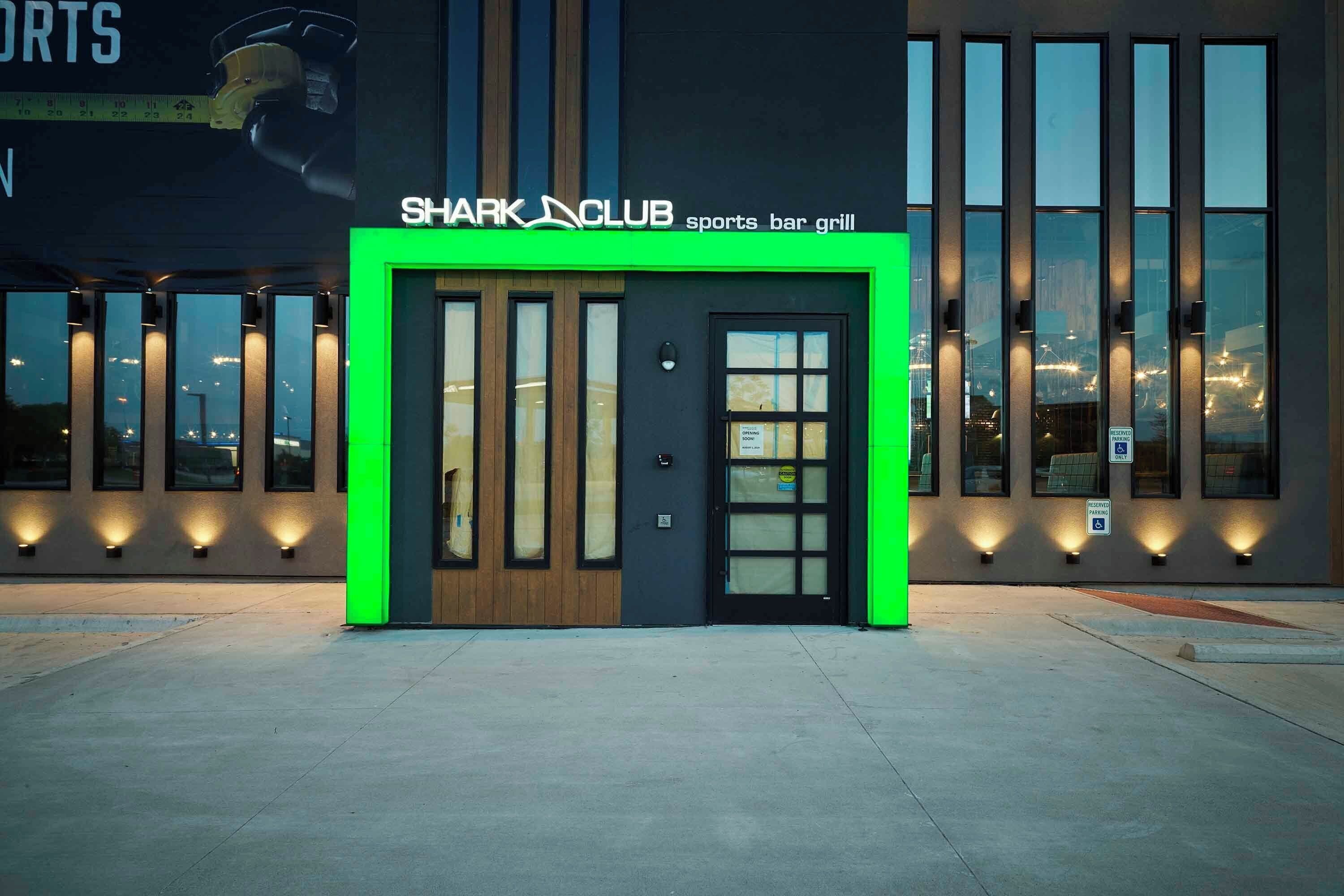 A sports bar and grill named Shark Club with a modern facade, large windows, and a green-lit entrance.