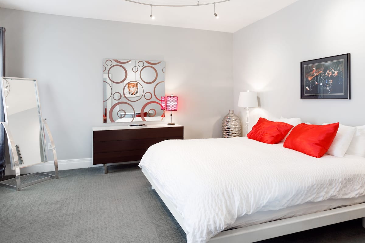 A modern bedroom with a white bed, red pillows, a dresser, a mirror, and a wall with abstract art.