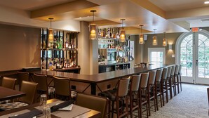 Una zona de bar con una barra larga, varios taburetes y una gran variedad de botellas y vasos en estanterías.