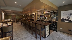 Una zona de bar con una barra alargada, varios taburetes y un estante bien surtido con botellas y vasos.