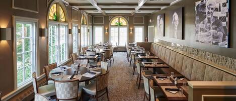 A spacious dining area with multiple tables and chairs, large windows, and artwork on the walls.