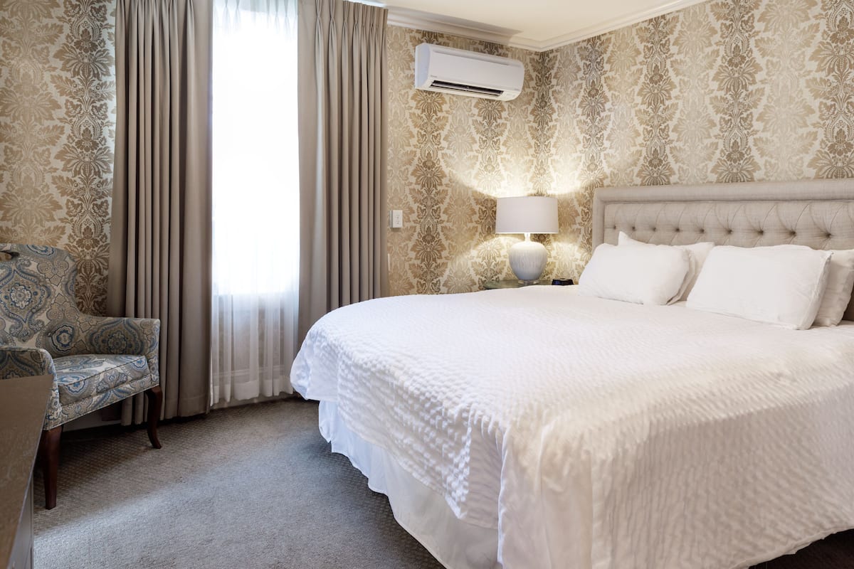 A hotel room with a large bed, a chair, an air conditioning unit, and a wall-mounted lamp.