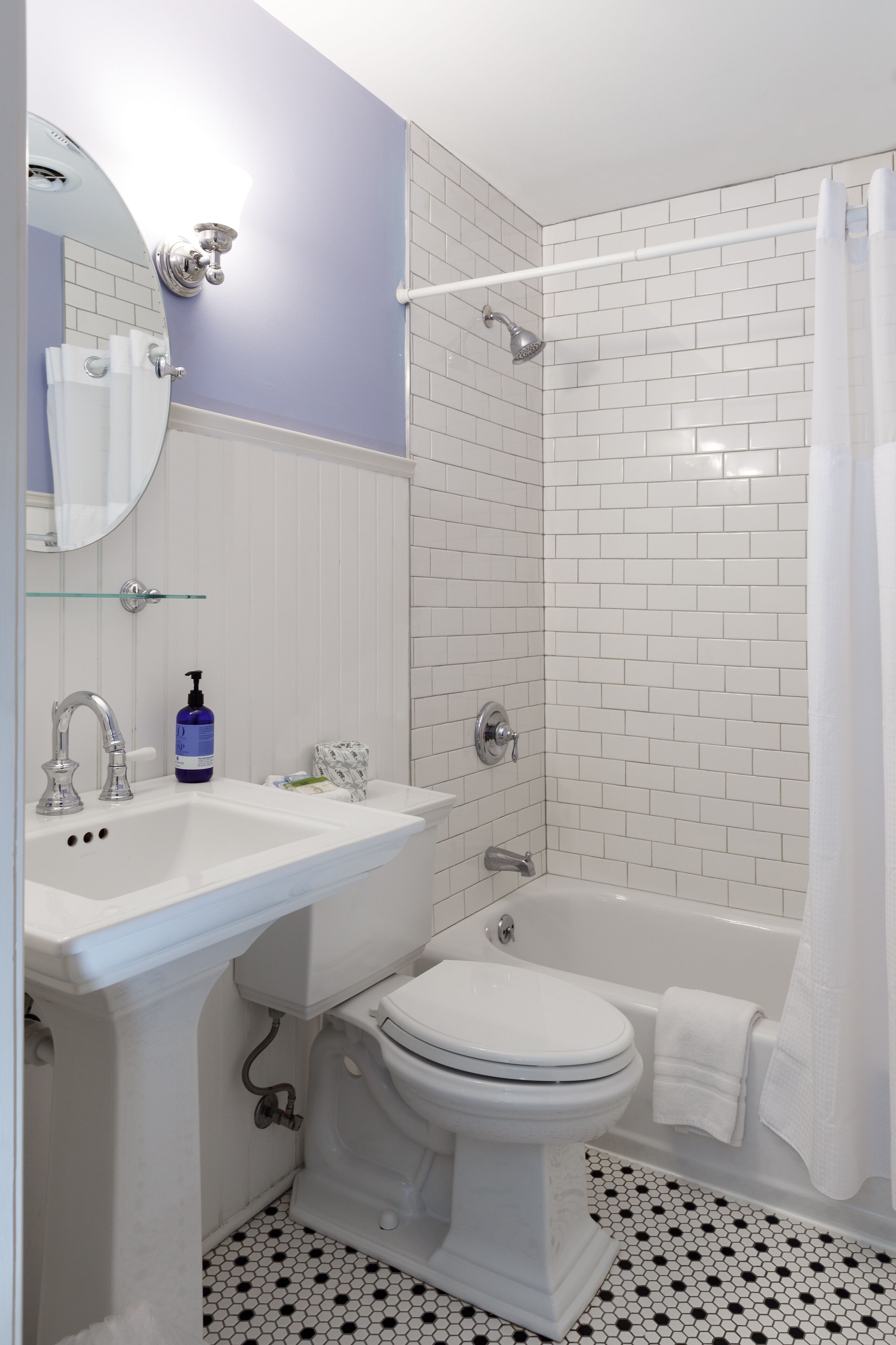 Un baño con lavamanos, inodoro y bañera blancos. Hay un estante de vidrio sobre el lavamanos, y se ve una cortina de ducha al fondo.