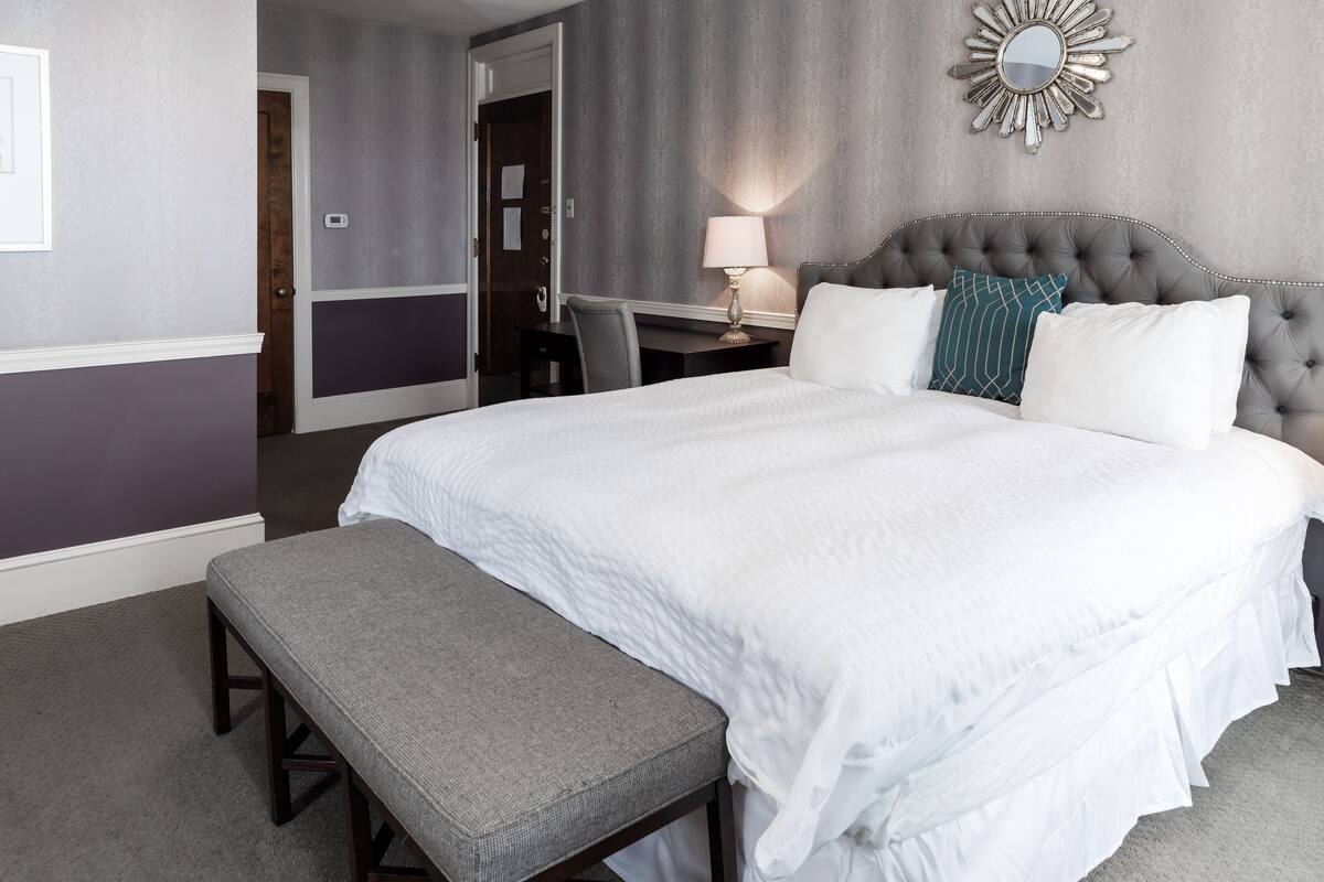 A neatly arranged hotel room with a large bed, a bedside table with a lamp, a chair, and a bench.