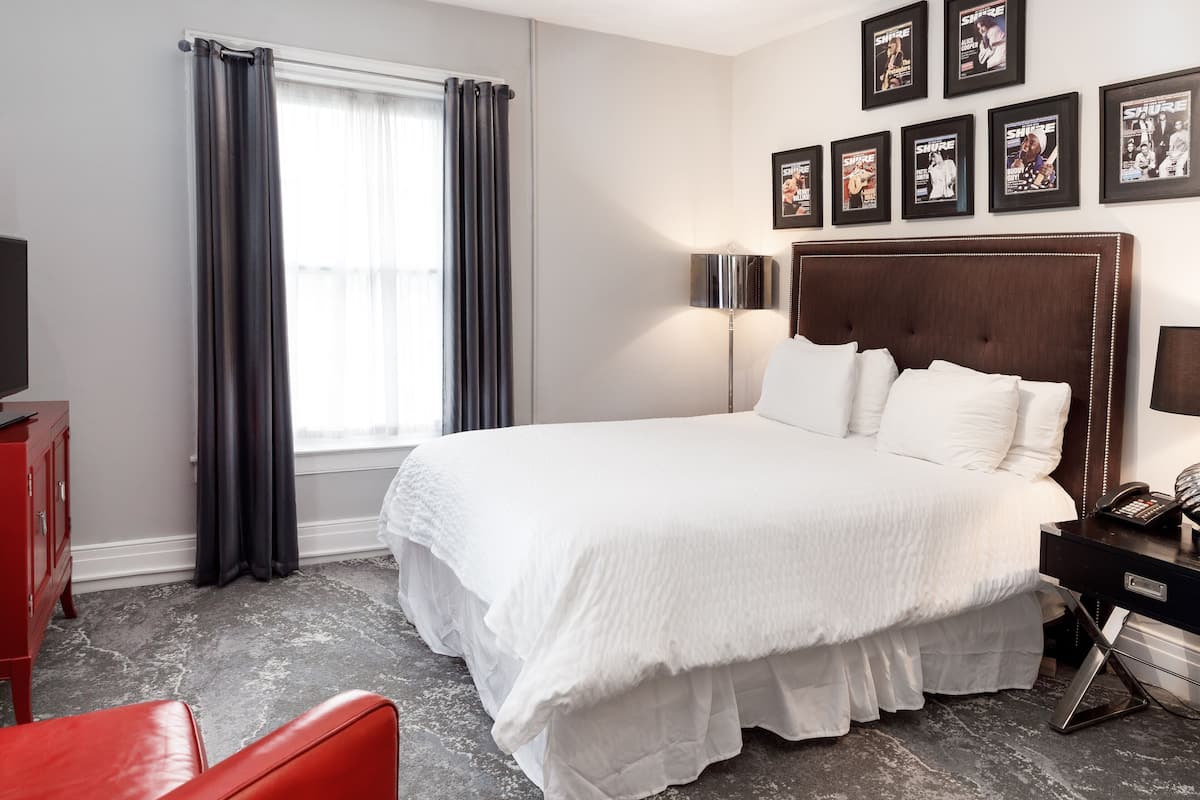 A hotel room with a large bed, a bedside table with a lamp, a red chair, and a window with curtains.