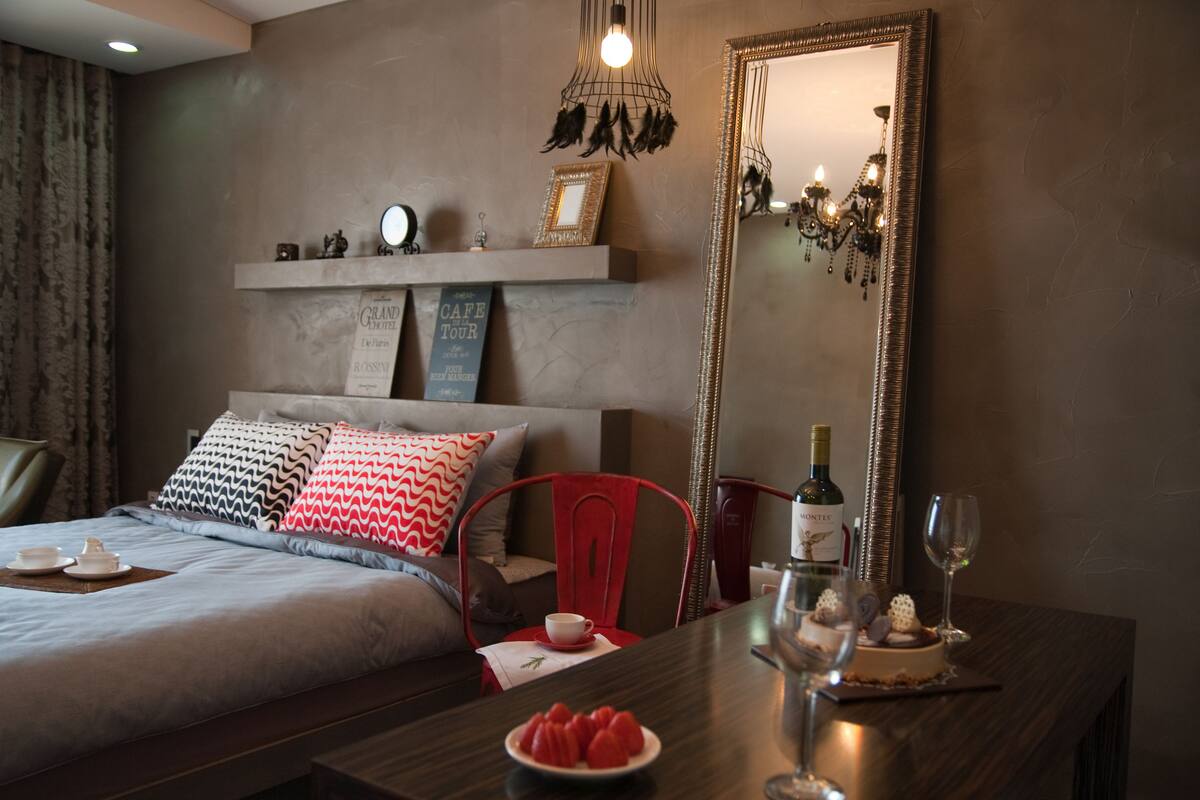 A hotel room with a bed, a dining table with a bottle of wine, a glass, and a plate of strawberries. There is a mirror on the wall and a chandelier above the table.