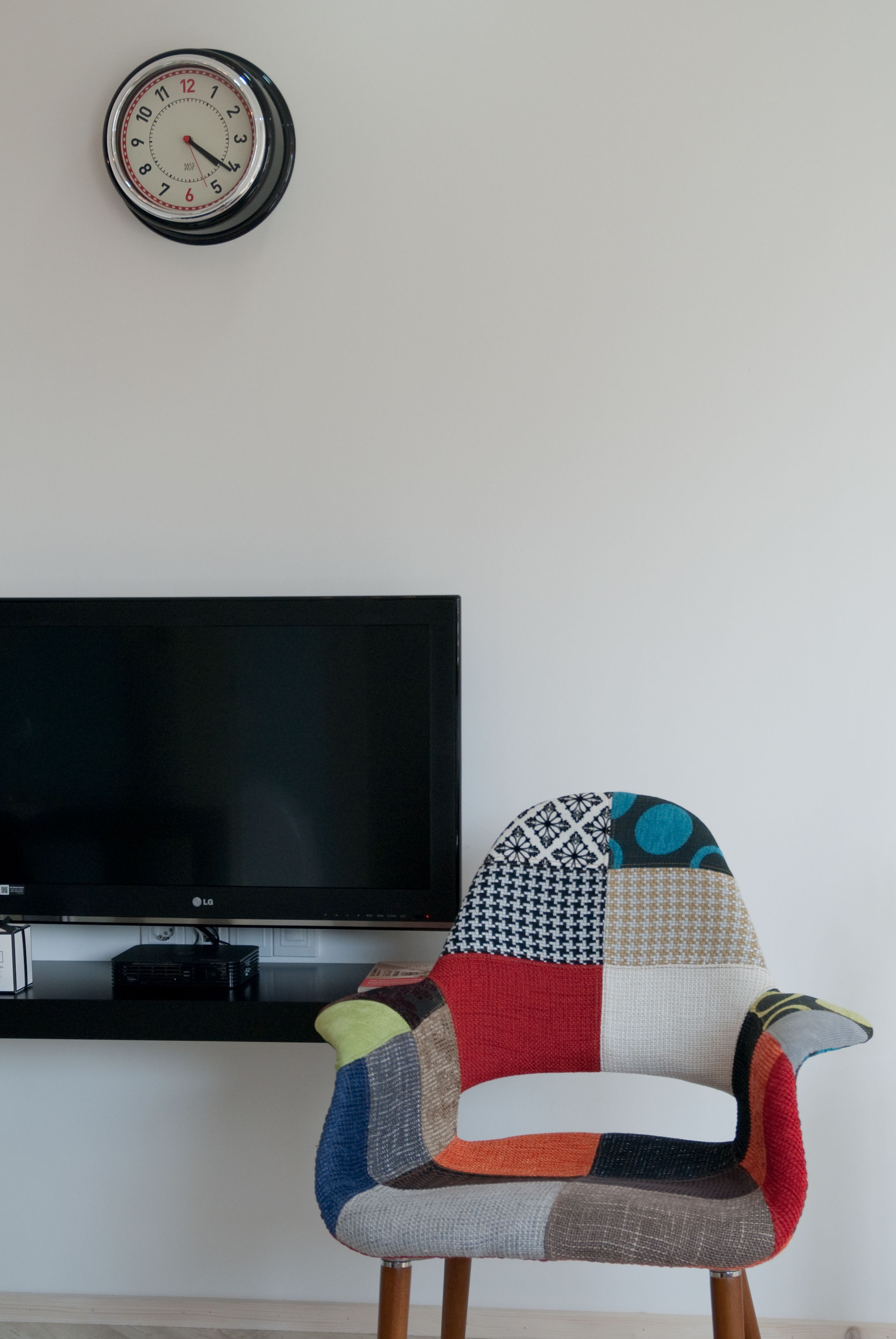A modern living room with a flat-screen TV, a colorful patchwork armchair, and a wall clock.
