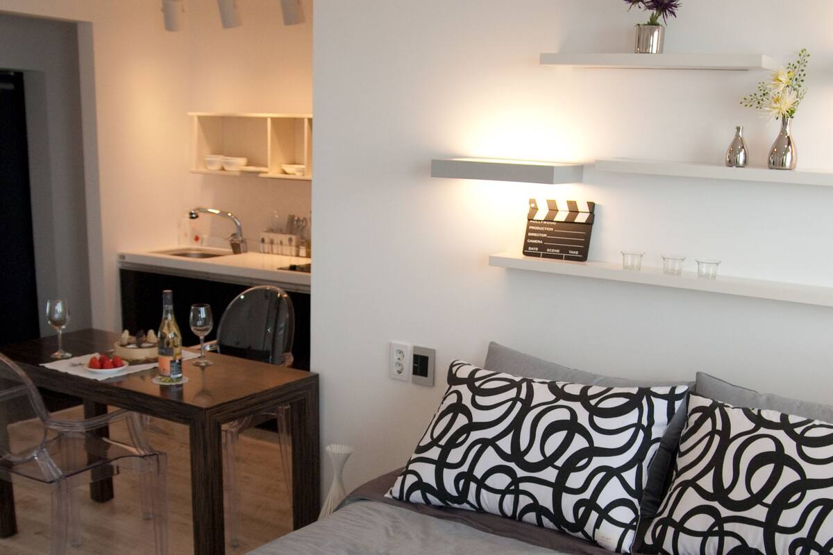 A modern hotel room with a neatly made bed, a dining table with chairs, and a kitchen area in the background.