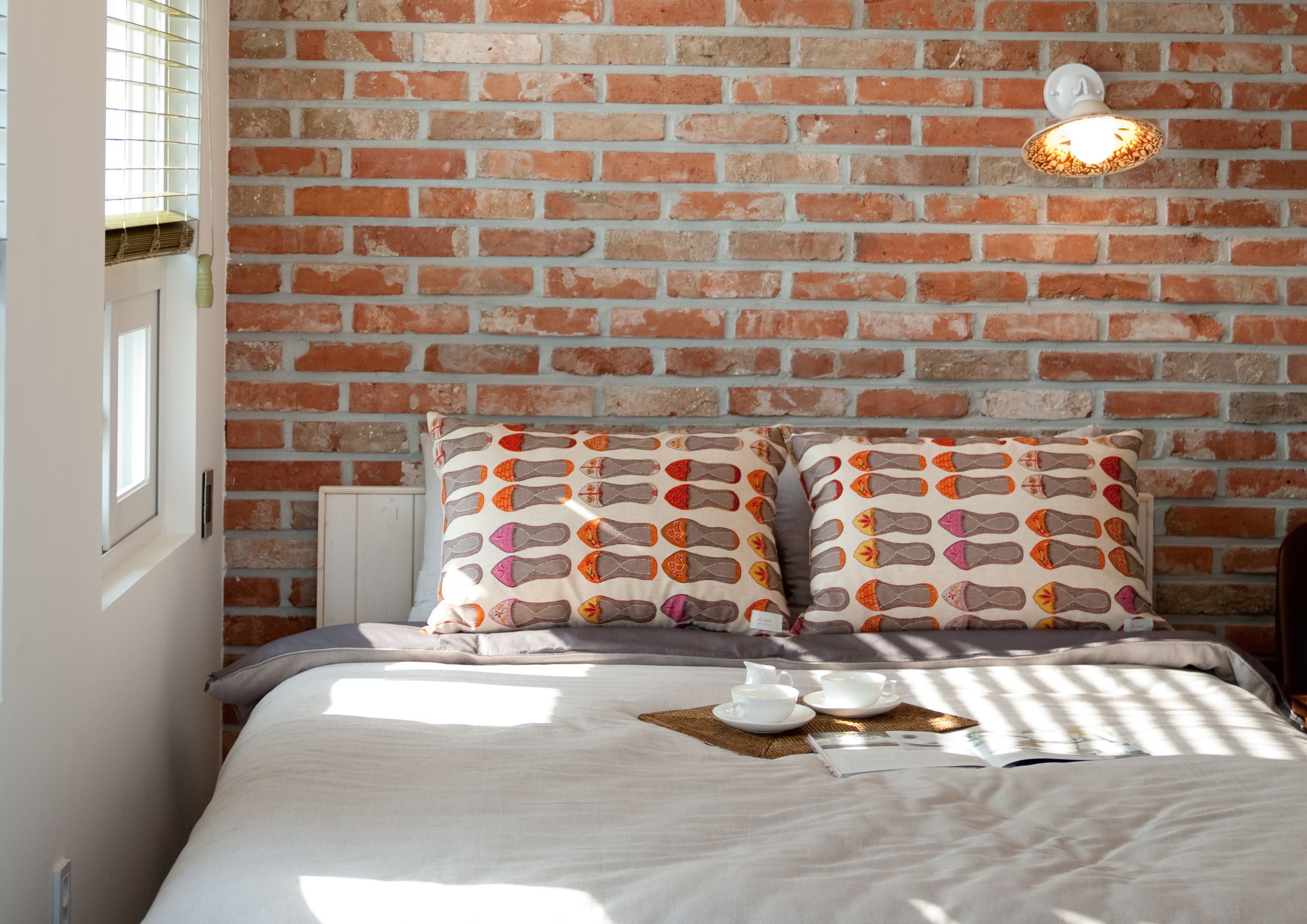 A bedroom with a brick wall, two patterned pillows, a bedside table with a lamp, and a breakfast setup on the bed.