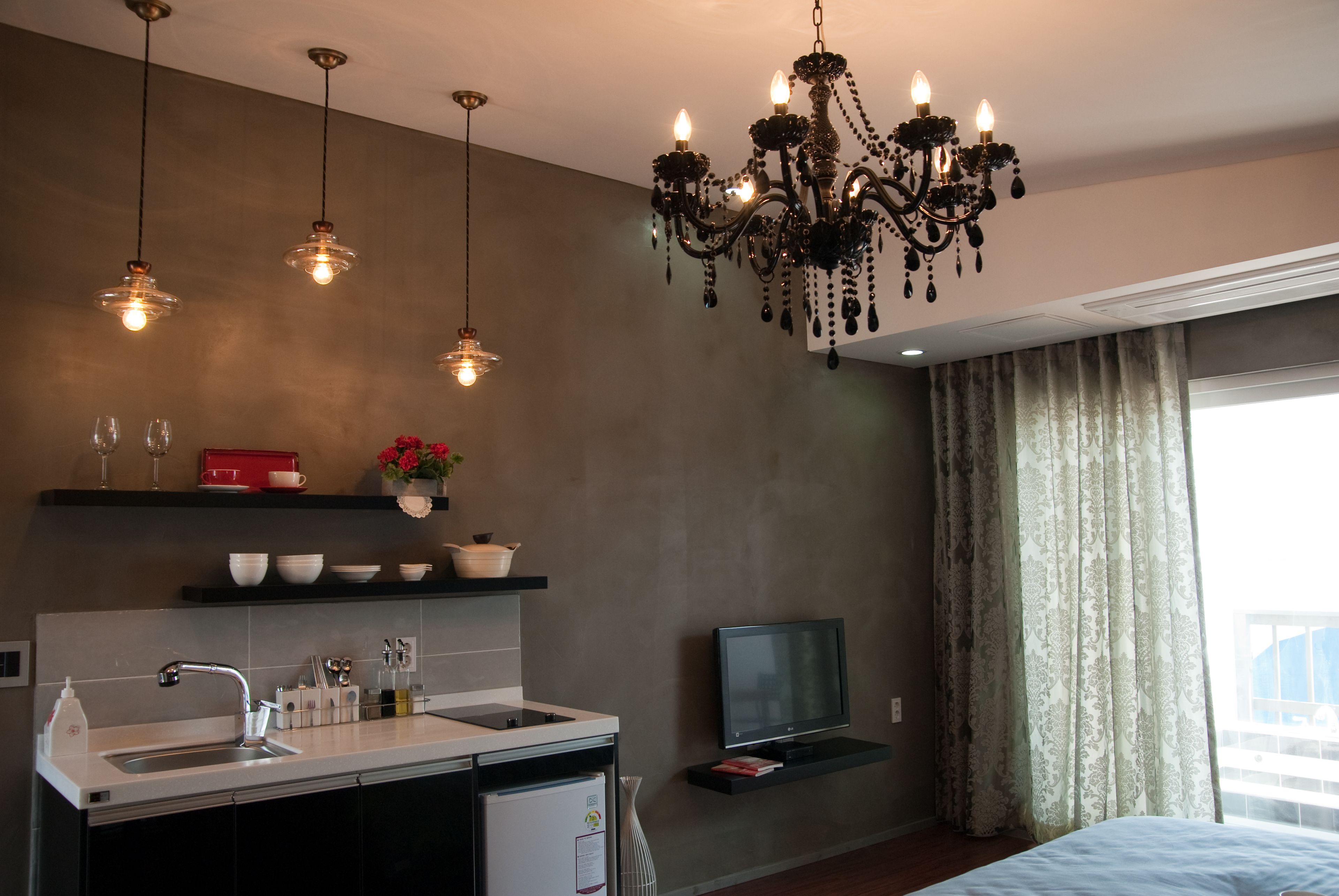 A modern kitchen with a sink, stove, and a flat-screen TV. There is a chandelier above the kitchen, and a bed with a window to the right.