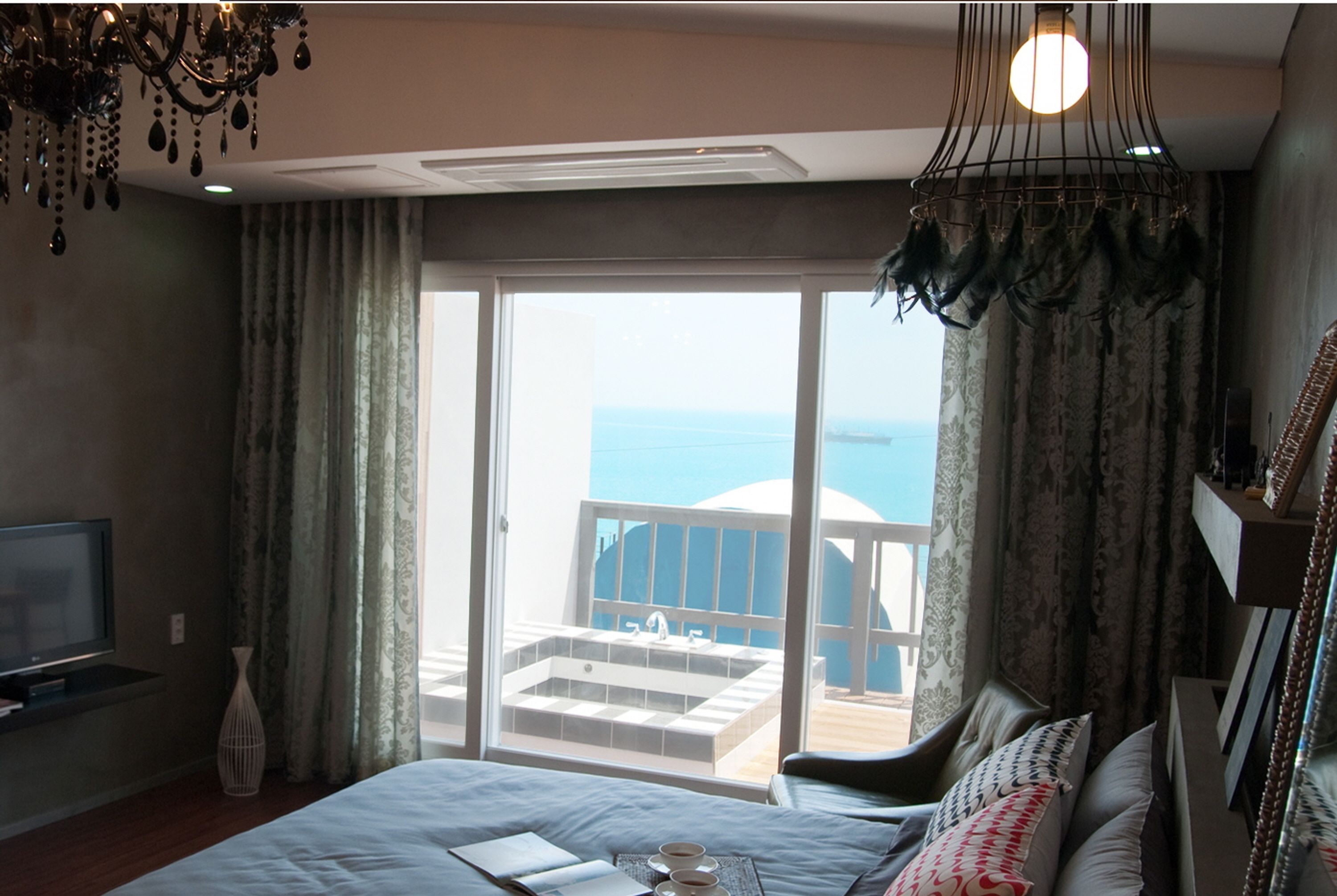 A hotel room with a bed, a television, a balcony with a view of the sea, and a chandelier.